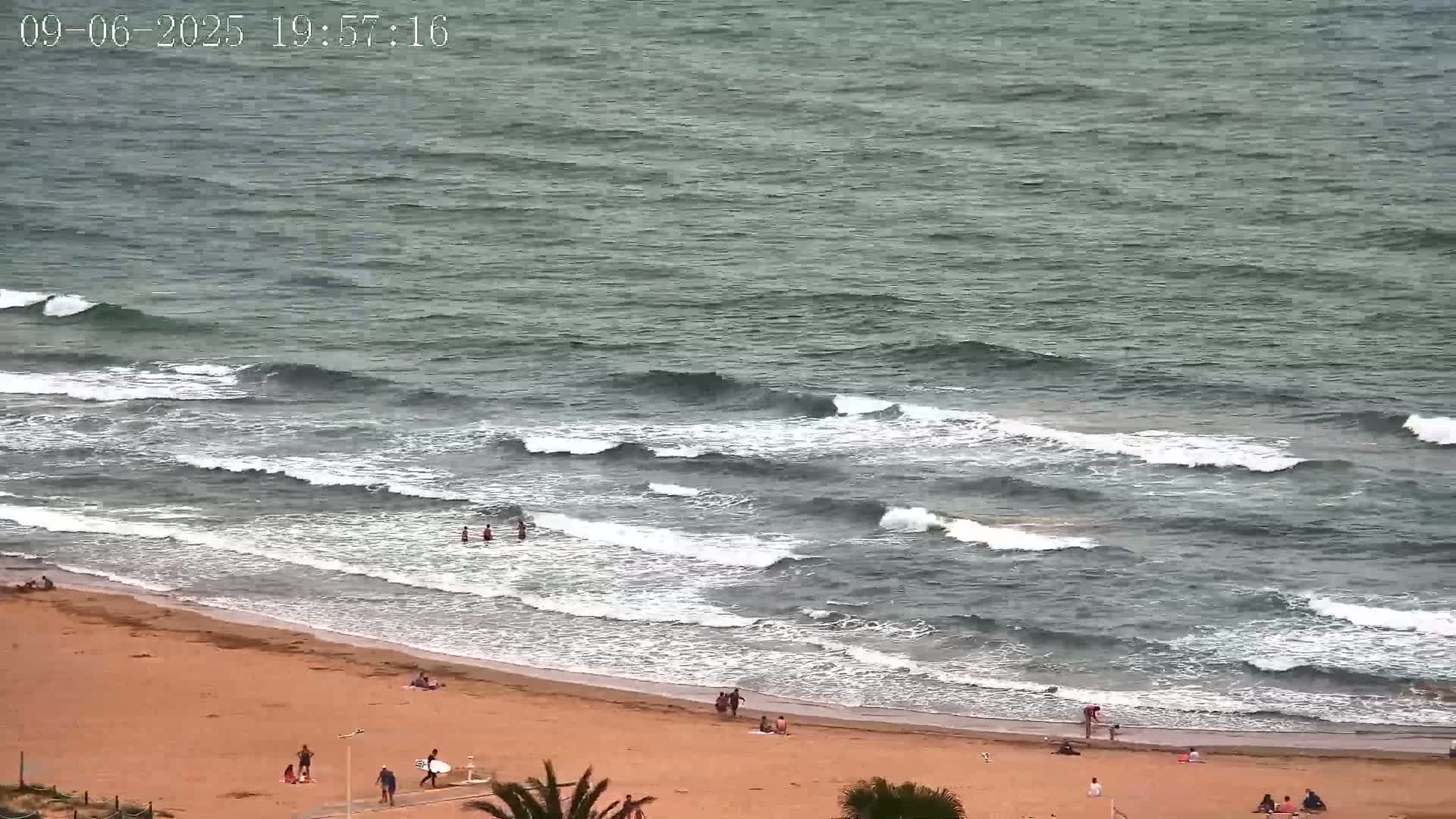 A sandy beach with a few people on it is met by a dark-grey sea with small waves breaking on the shore.