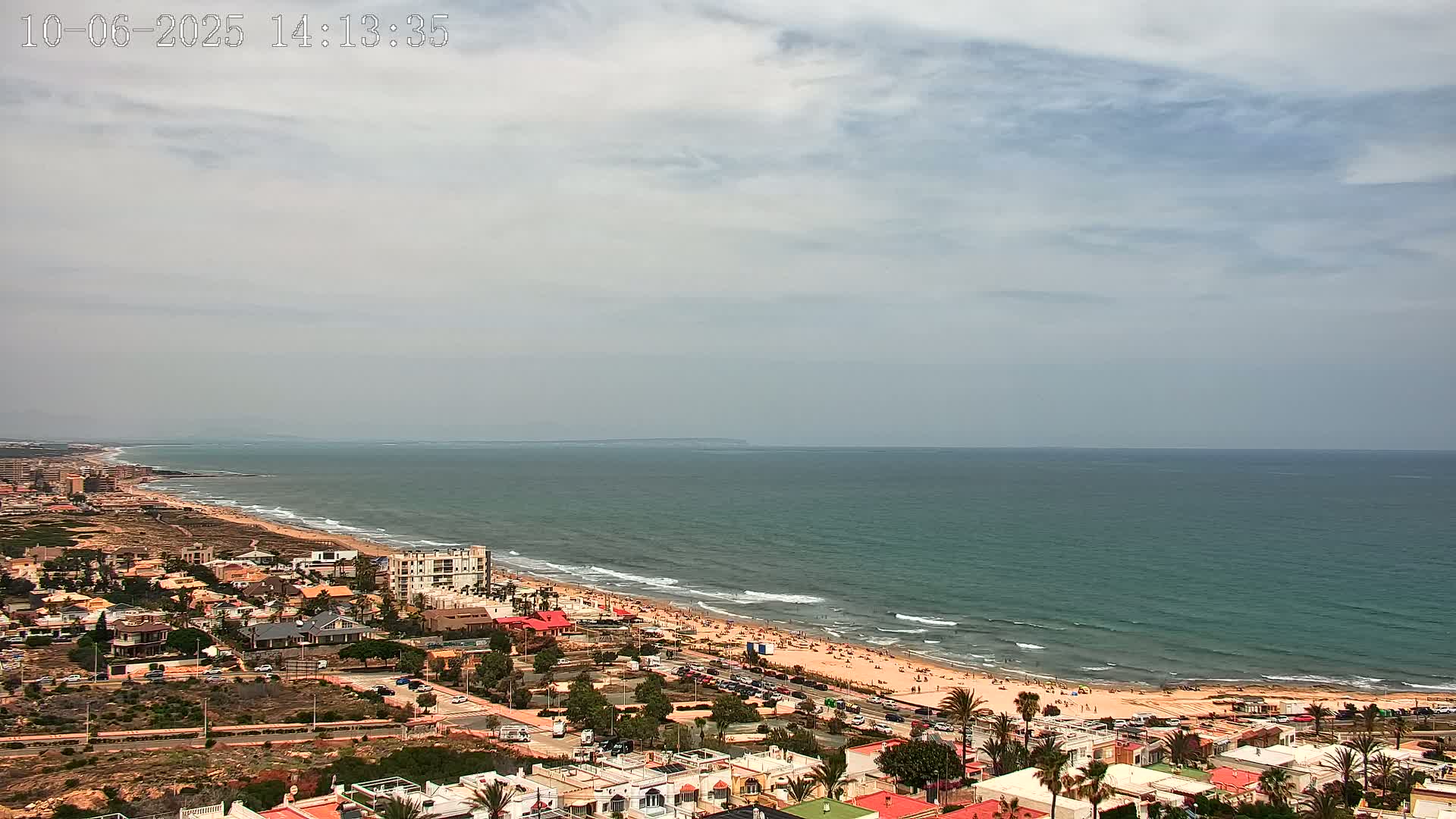 An aerial view shows a coastal town with a sandy beach and calm ocean under a partly cloudy sky.