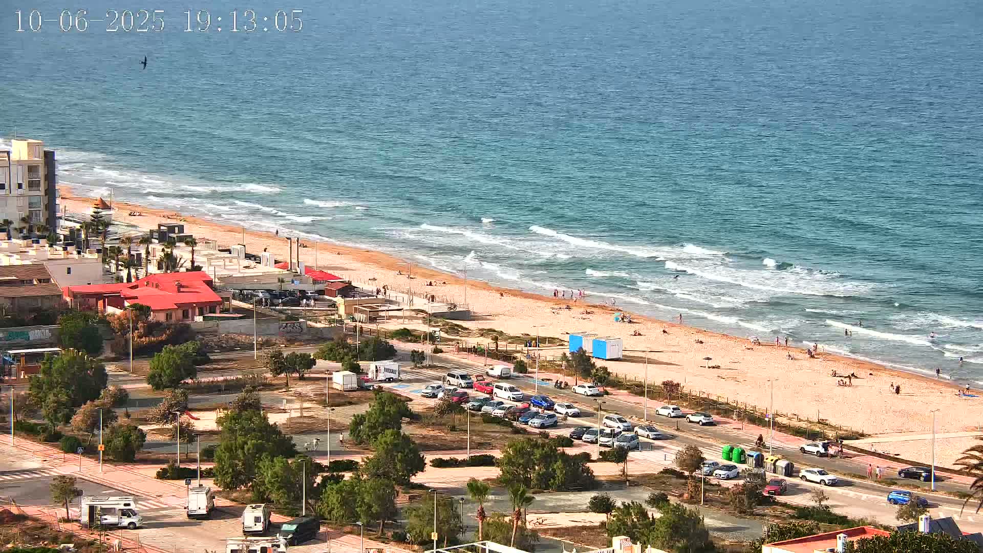 A sandy beach with people sunbathing and swimming in the ocean under a sunny sky, next to a parking lot and buildings.