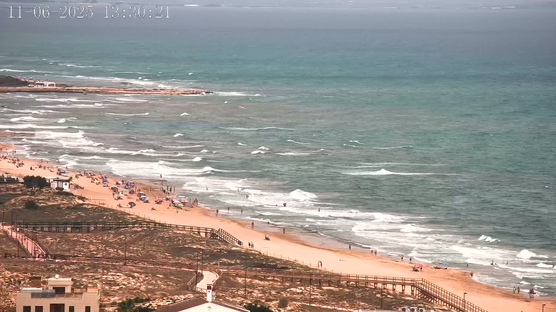 A sandy beach with many people on it is bordered by a boardwalk and dune grasses, with moderate waves breaking on the shore under a partly cloudy sky.