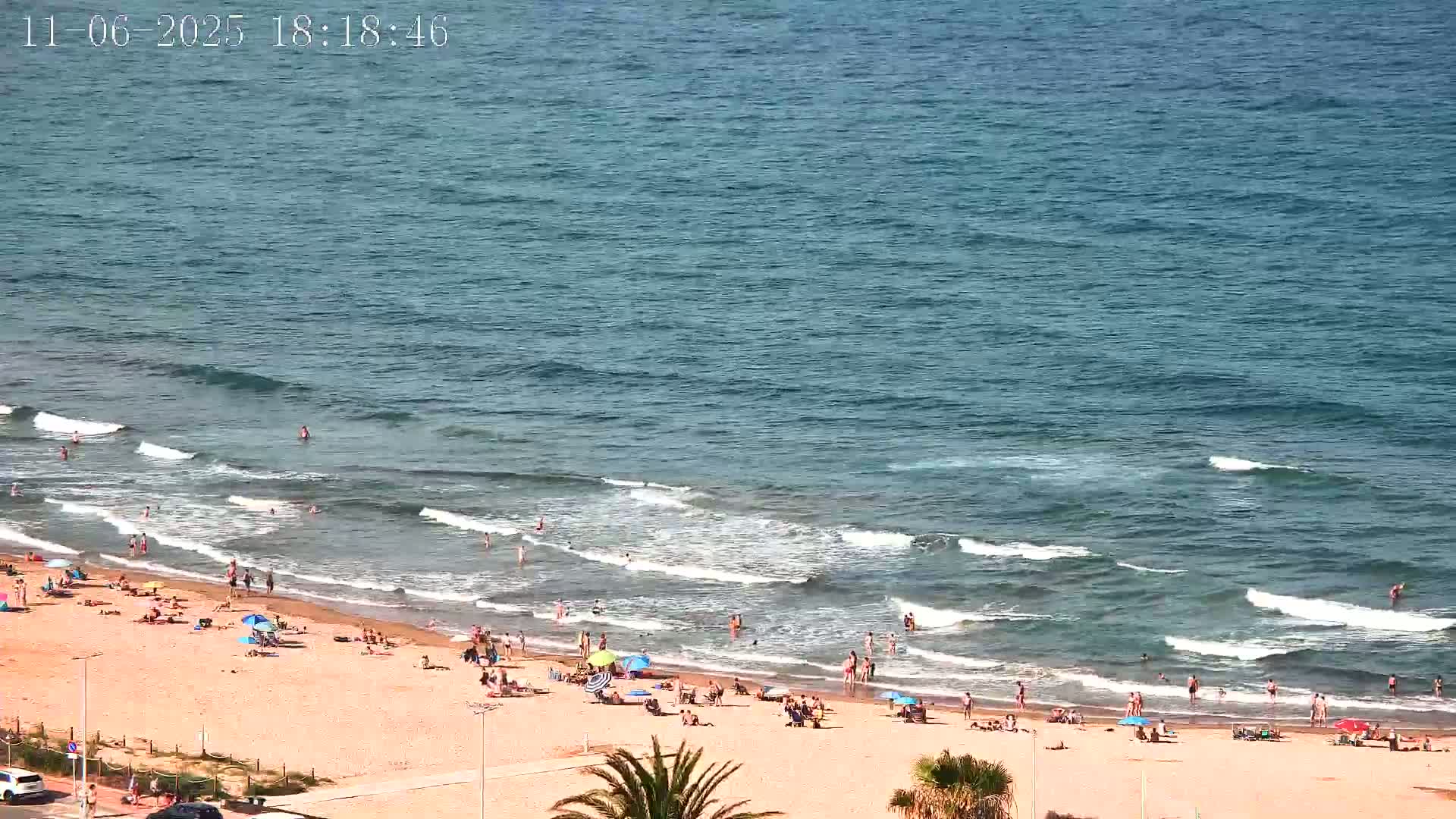 A sandy beach with numerous people swimming, sunbathing, and relaxing under umbrellas, bordered by a calm ocean with small waves under a sunny sky.