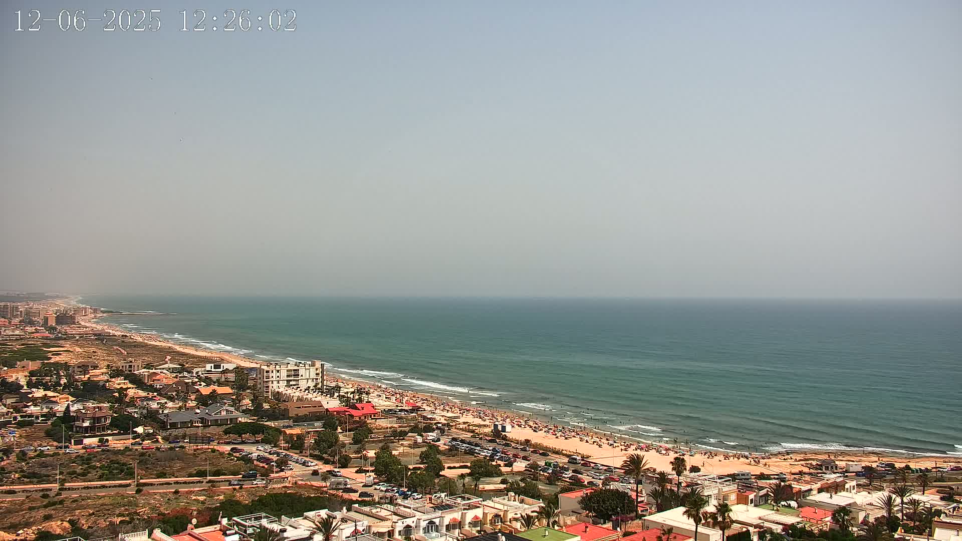 A hazy, sunny day shows a long sandy beach crowded with people, next to a coastal town, all overlooking a teal-colored ocean.