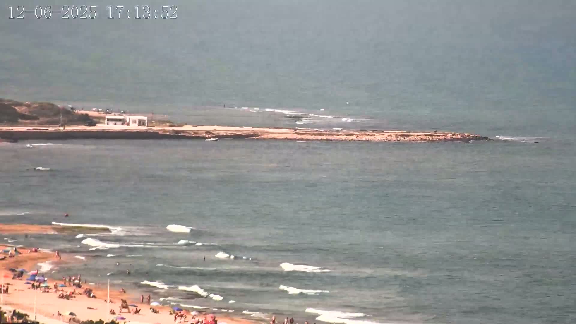 A hazy, sunny day at a beach shows a sandy shore with scattered people, gentle waves, and a small land formation extending into the ocean.