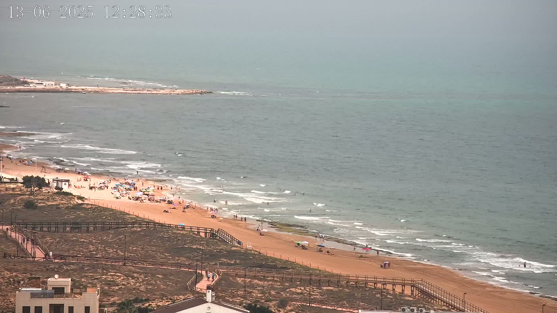 A hazy day at a beach shows a sandy shore with many people, gentle waves, and a boardwalk leading to the ocean.