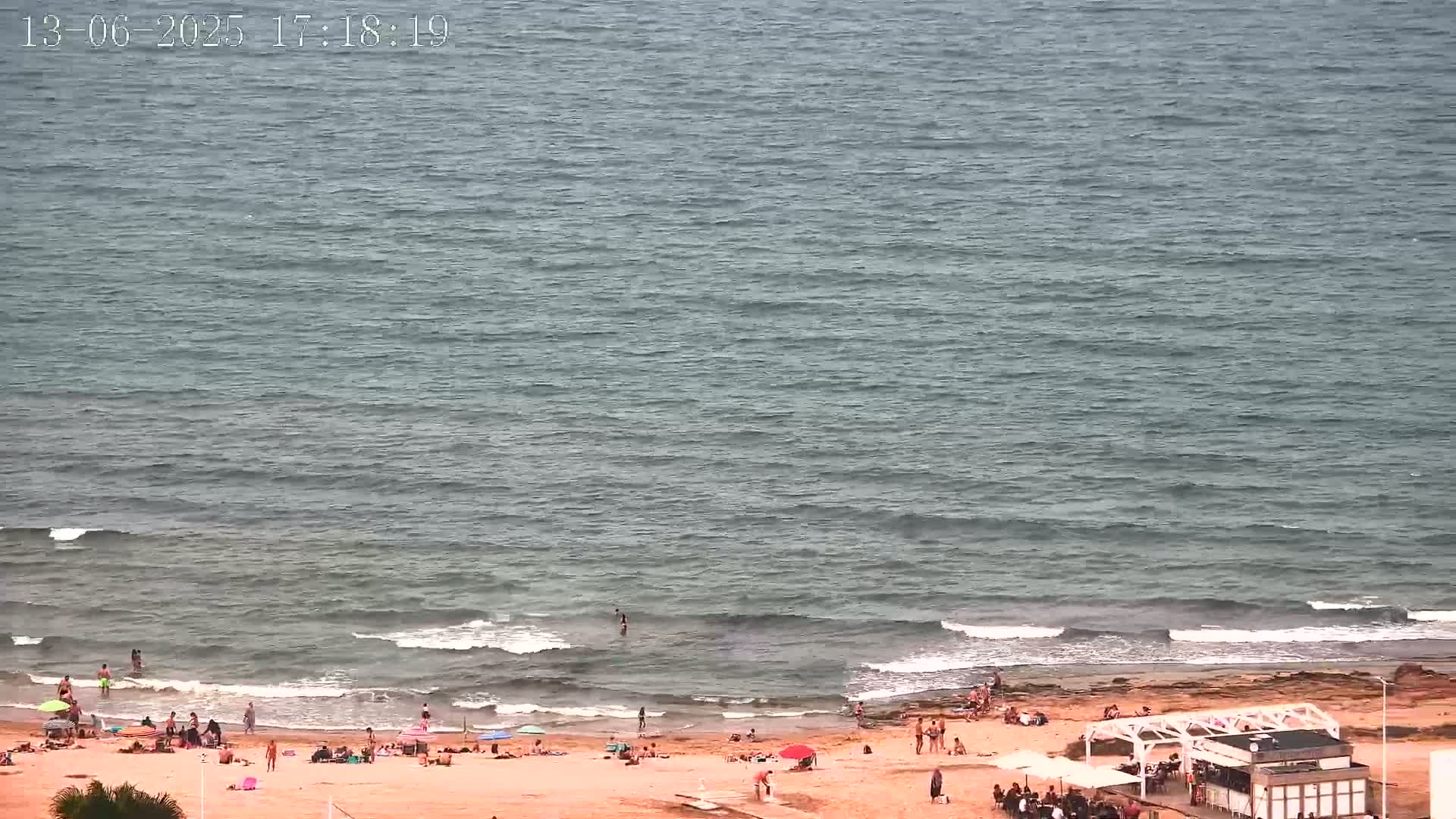 A sandy beach with people sunbathing and swimming in the ocean under a mostly cloudy sky.