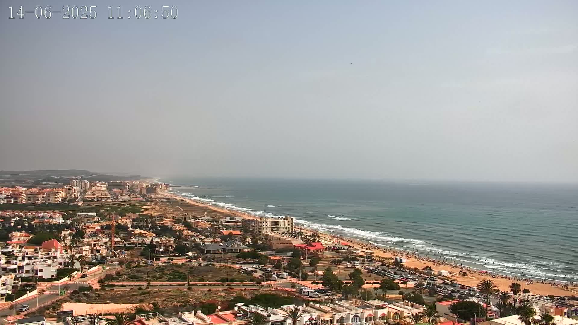 An aerial view shows a hazy coastline with a sandy beach, ocean waves, and a town of low-rise buildings extending inland.