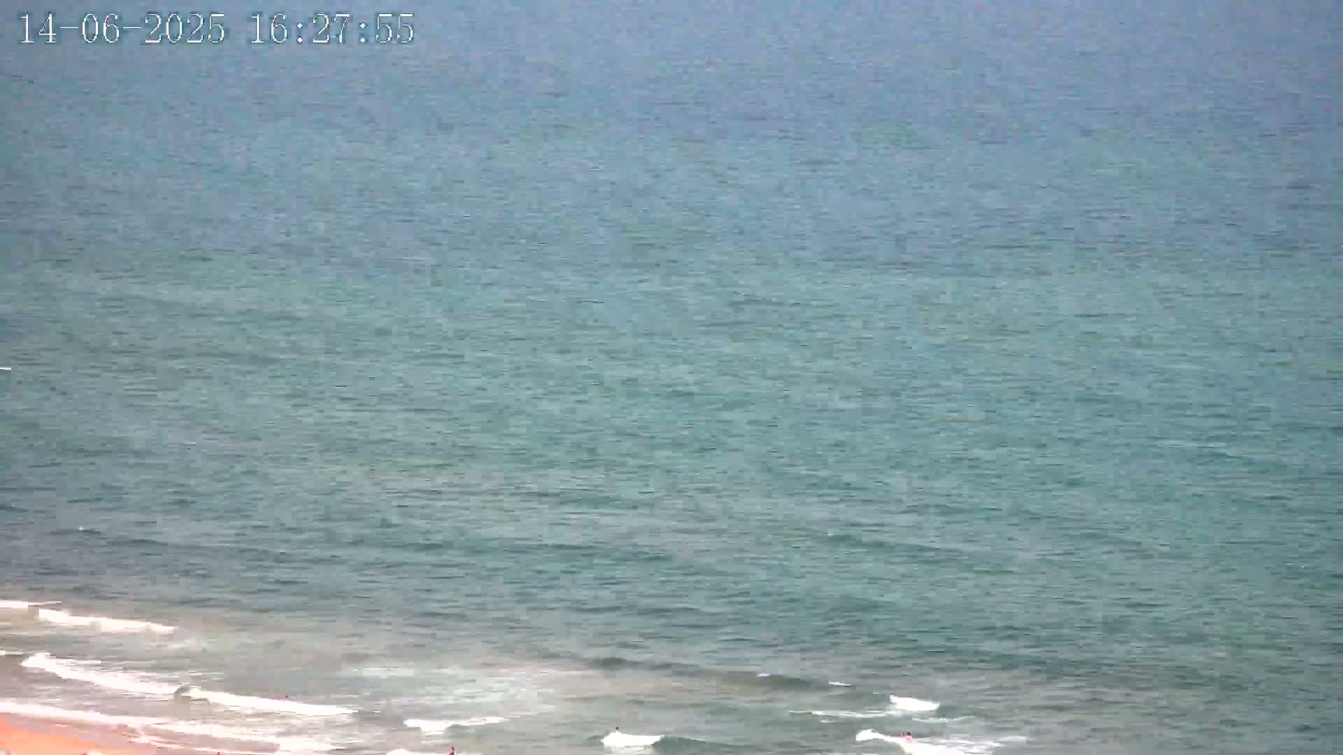 A mostly calm ocean with small waves breaking on a sandy beach under a cloudy sky.