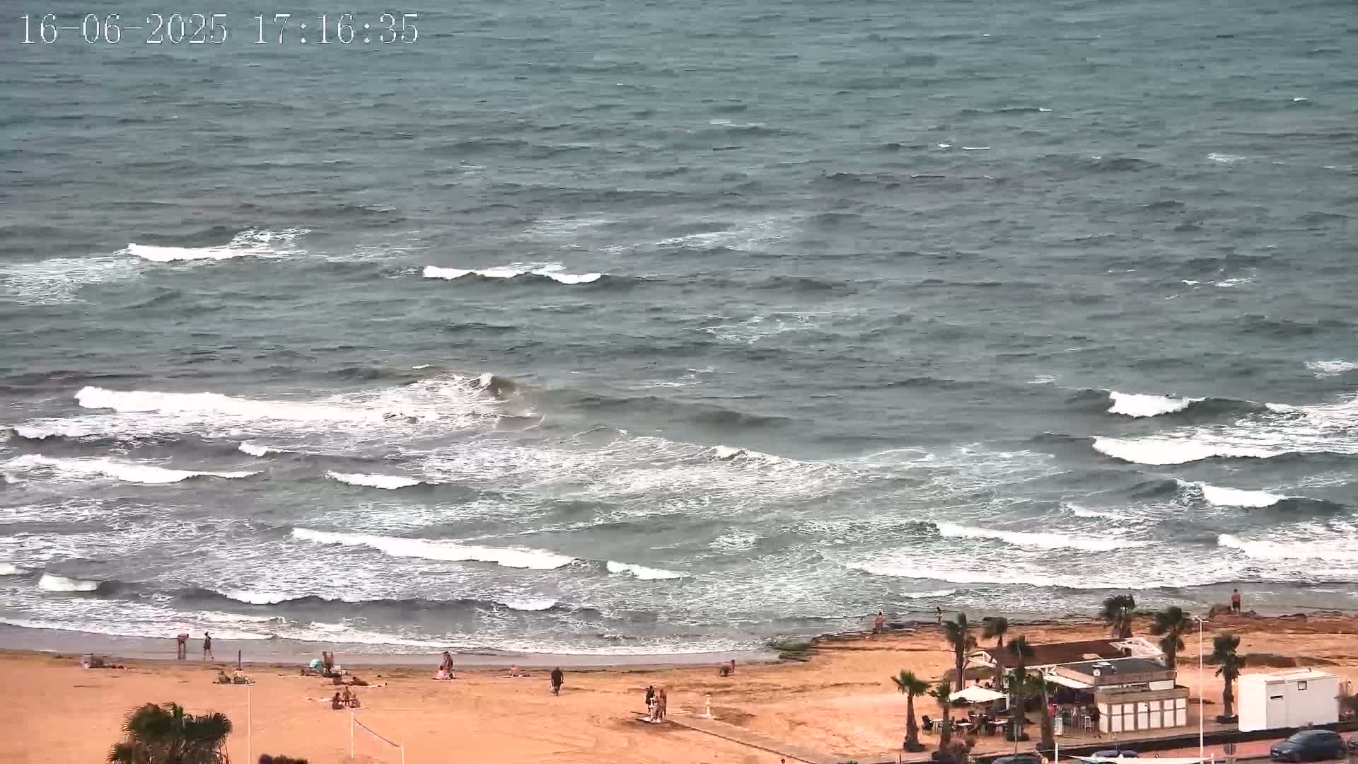 A sandy beach with a few people on it is bordered by a sea with moderately sized waves under a somewhat overcast sky.