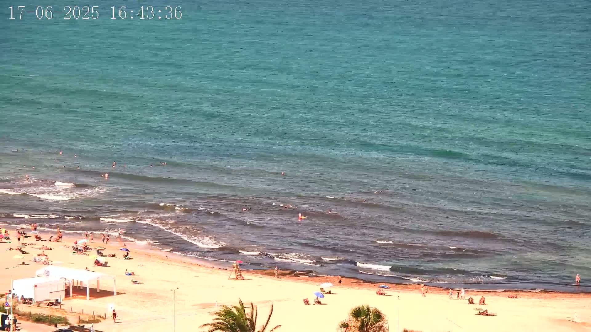 A sandy beach with numerous people sunbathing and swimming in a teal ocean under sunny skies.