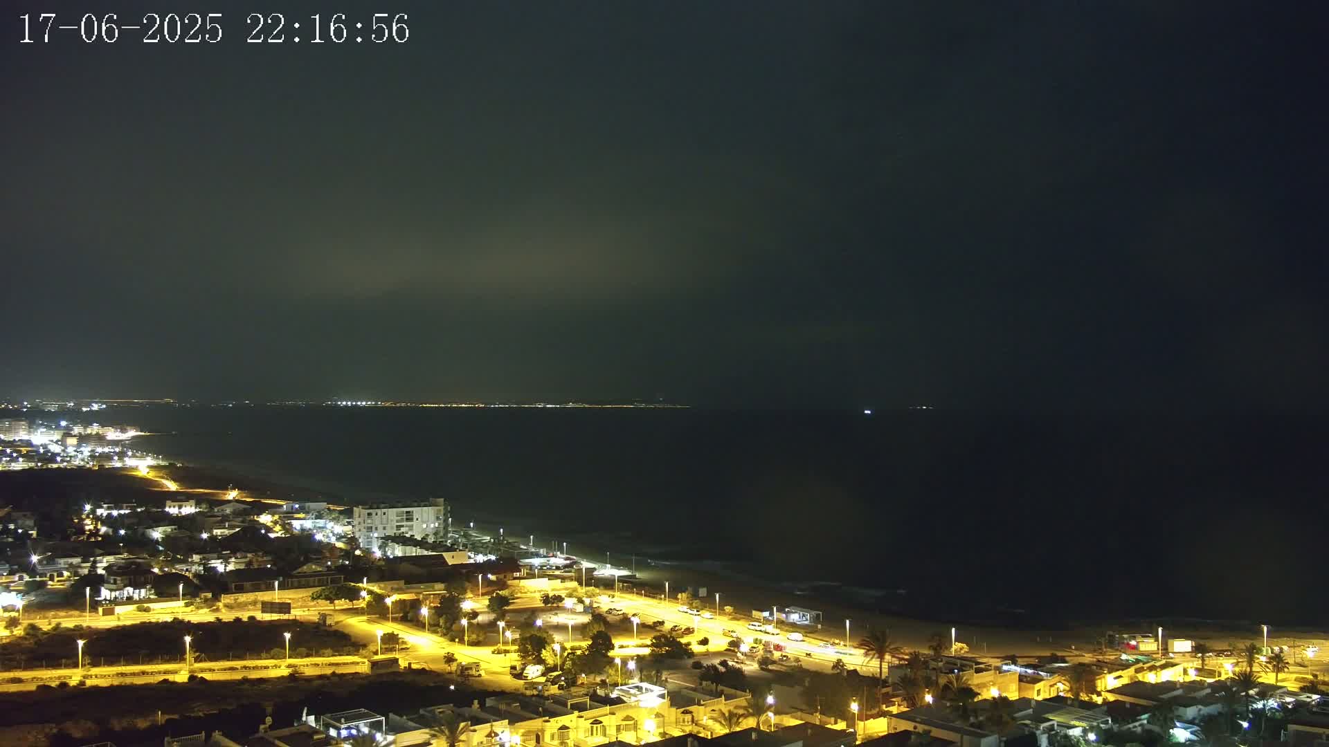 A night-time view of a coastal town illuminated by streetlights, overlooking a dark sea under a mostly clear, dark sky.