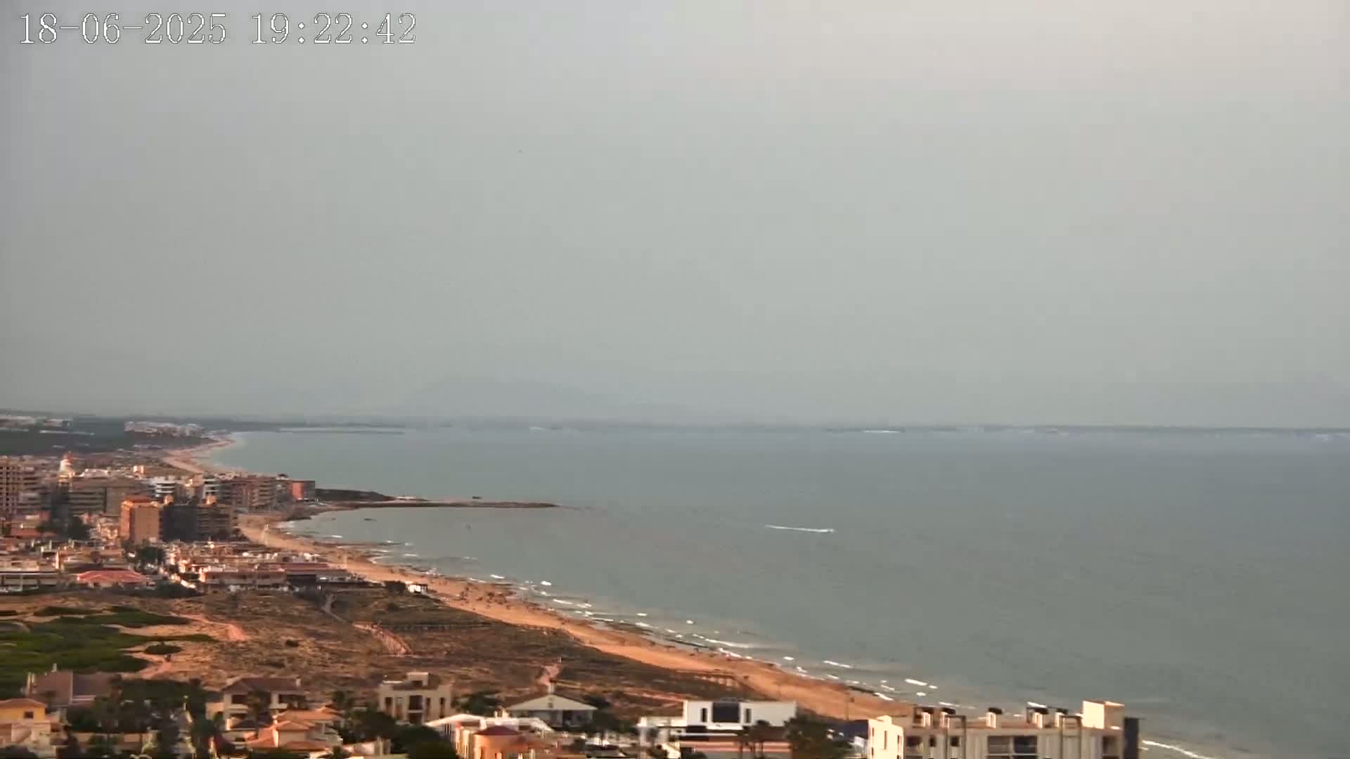 A hazy coastal town overlooks a calm, gray sea with a sandy beach.