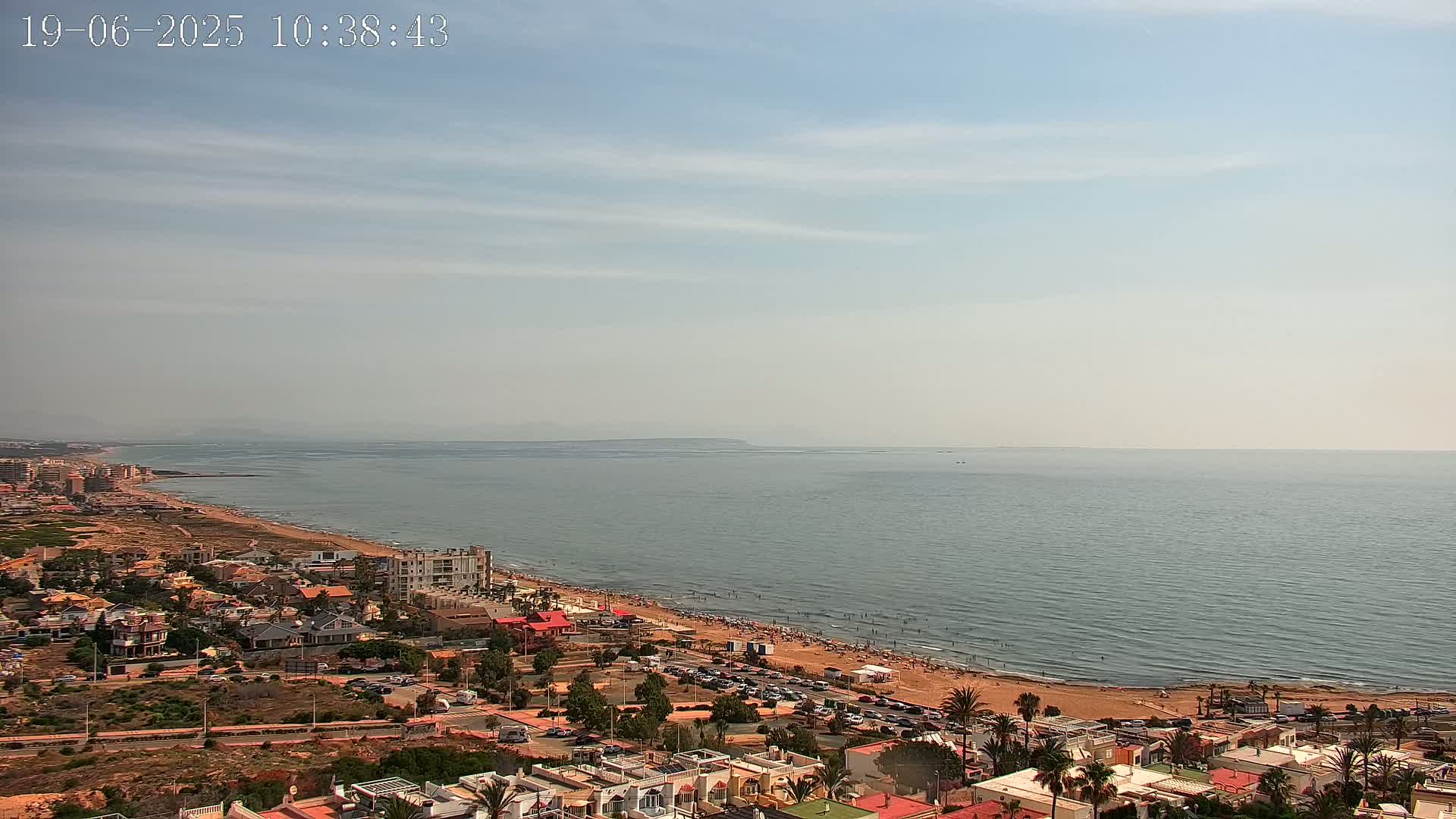 A mostly clear day shows a sandy beach with people swimming and sunbathing, next to a coastal town overlooking a calm sea and distant land.