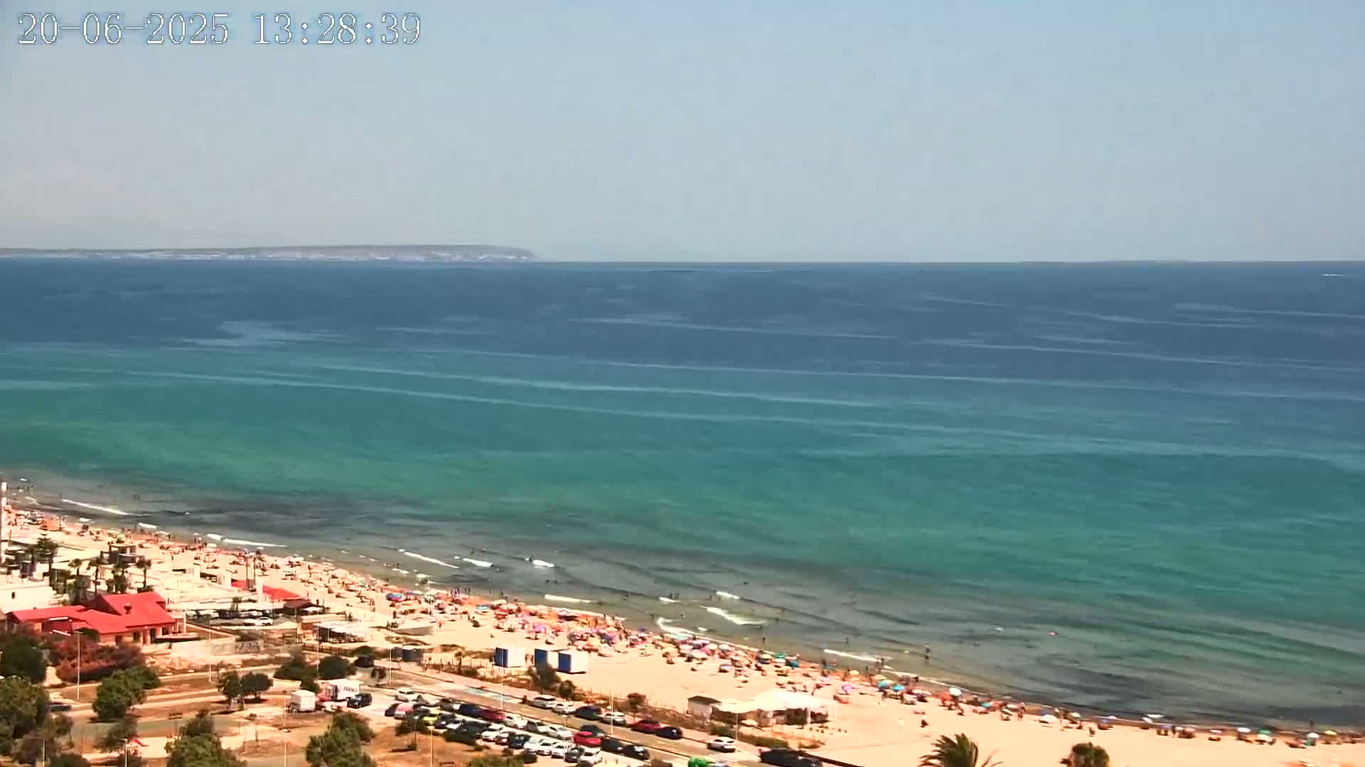 A crowded beach with turquoise water stretches to a distant landmass under a clear sky.
