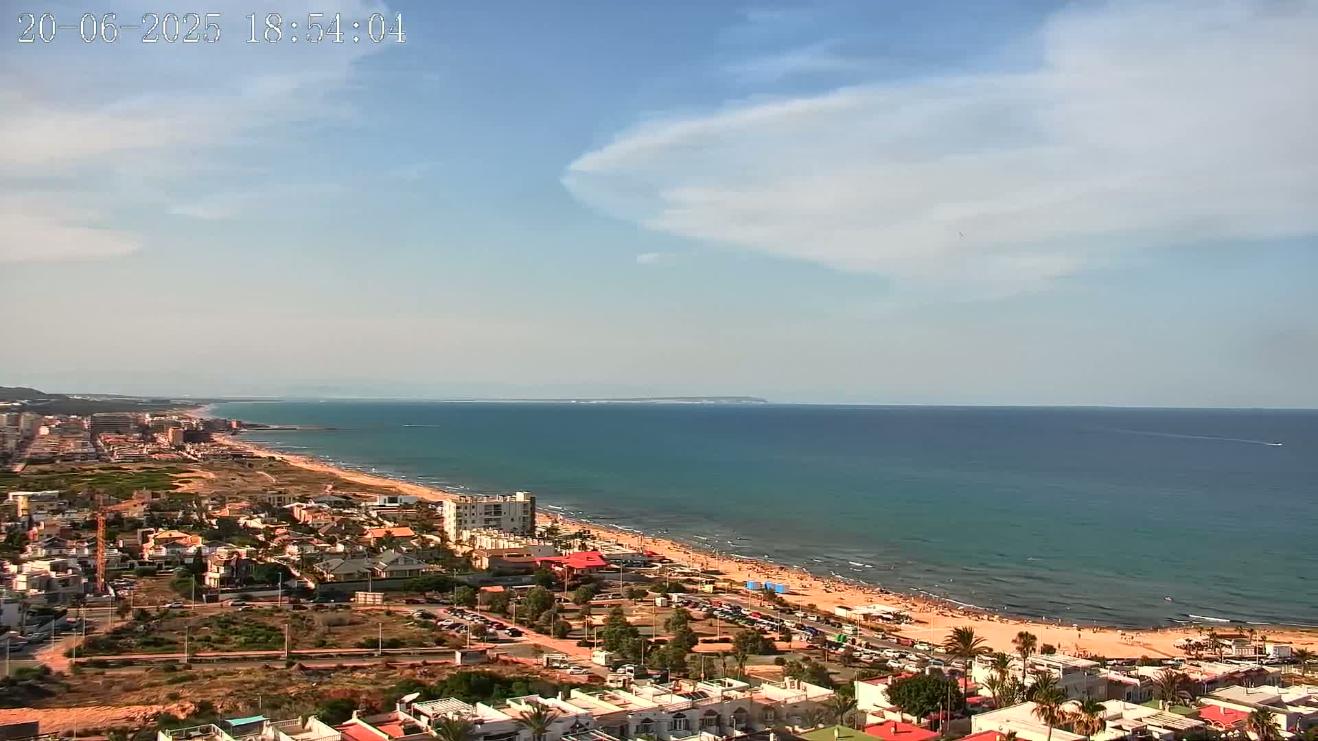 An aerial view shows a partly cloudy, sunny day overlooking a beach town and ocean with people enjoying the beach.