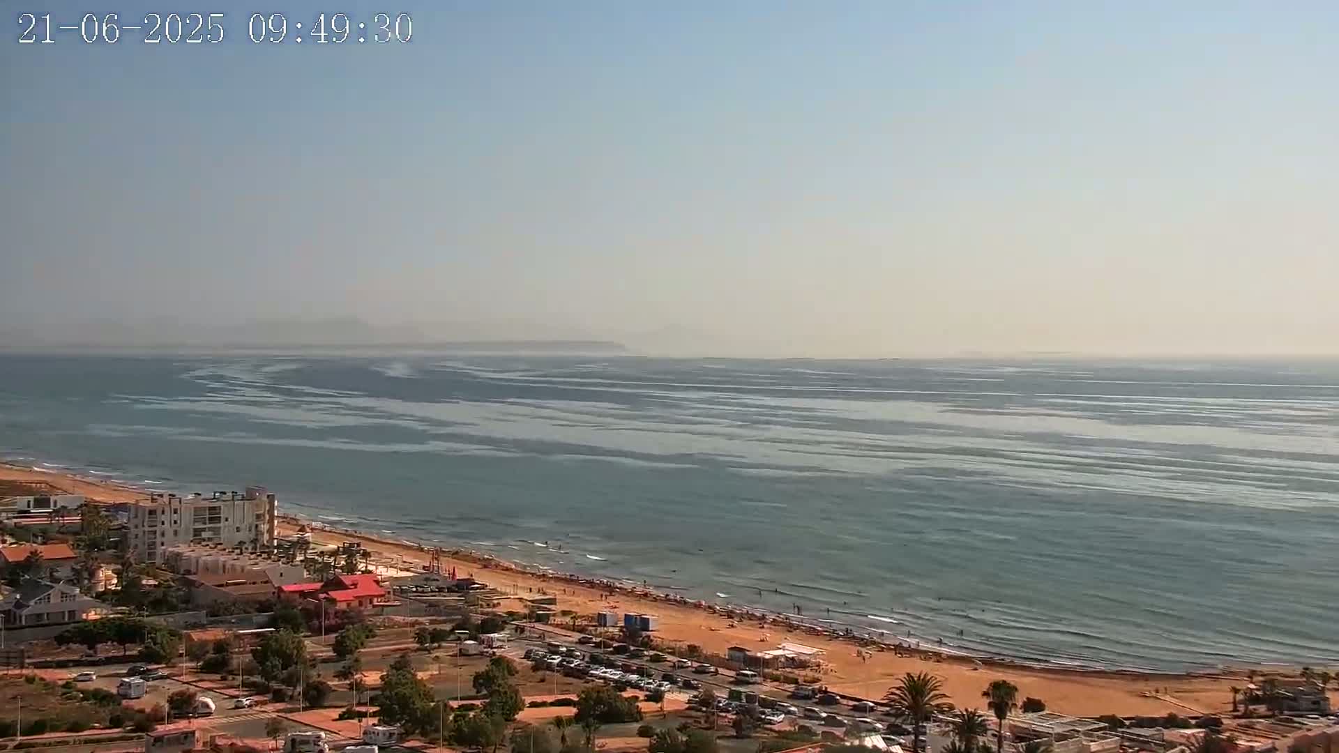 A hazy, sunny day shows a long sandy beach with a calm ocean, buildings, and parked cars along the coast.