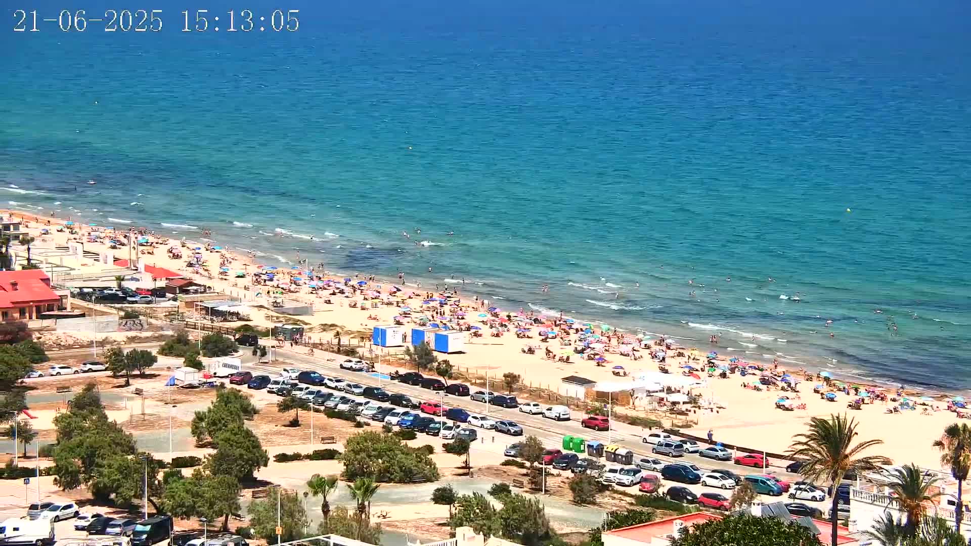 A crowded sandy beach with turquoise water under a clear blue sky on a sunny day, features numerous parked cars and beach umbrellas.