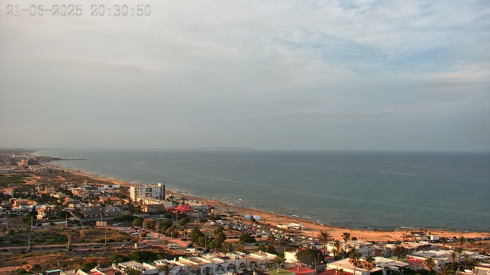 An elevated view shows a coastal town under a partly cloudy sky, with a long sandy beach and calm ocean extending to the horizon.
