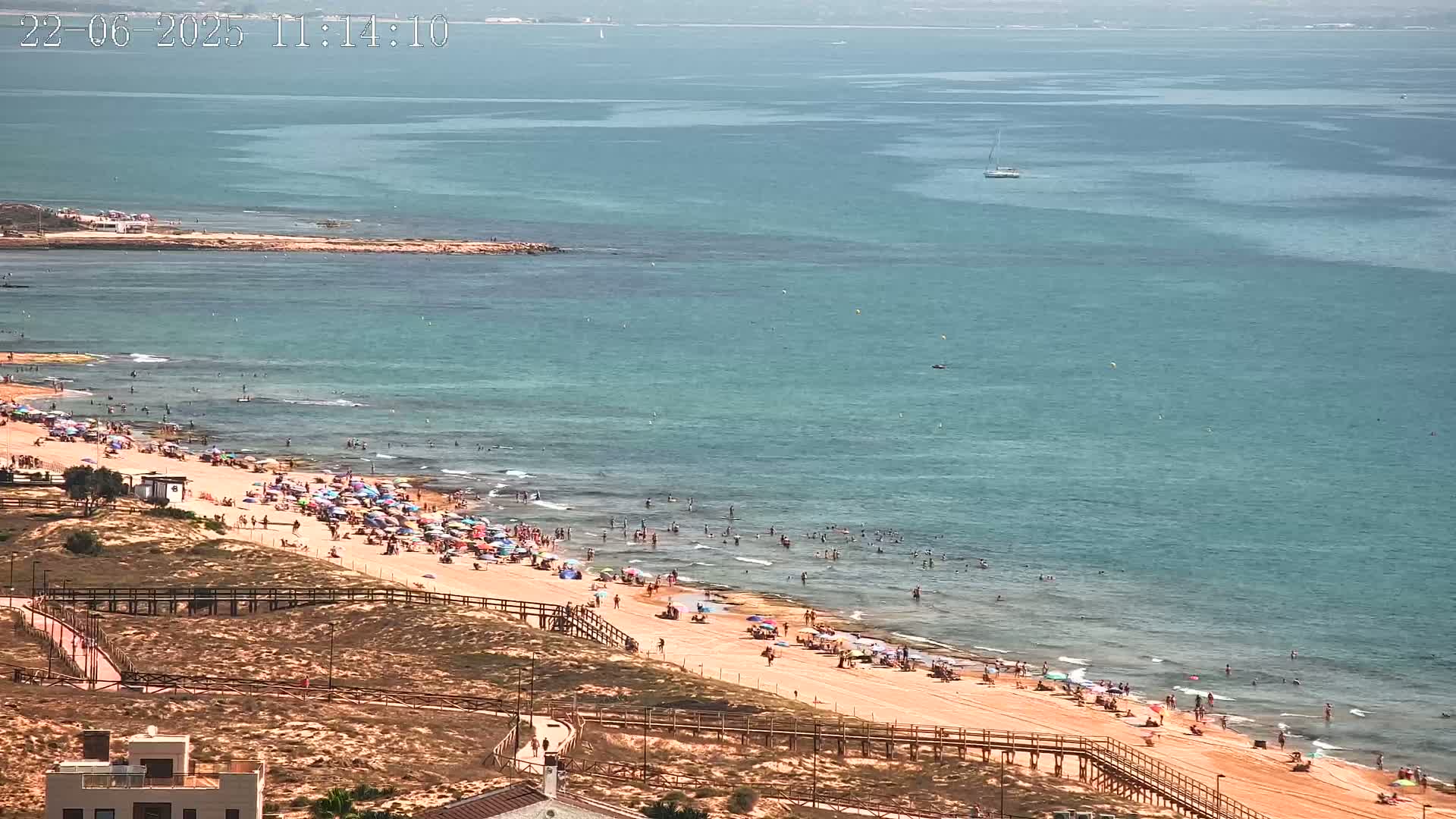 A sandy beach crowded with people under umbrellas, next to calm turquoise water, on a sunny day.