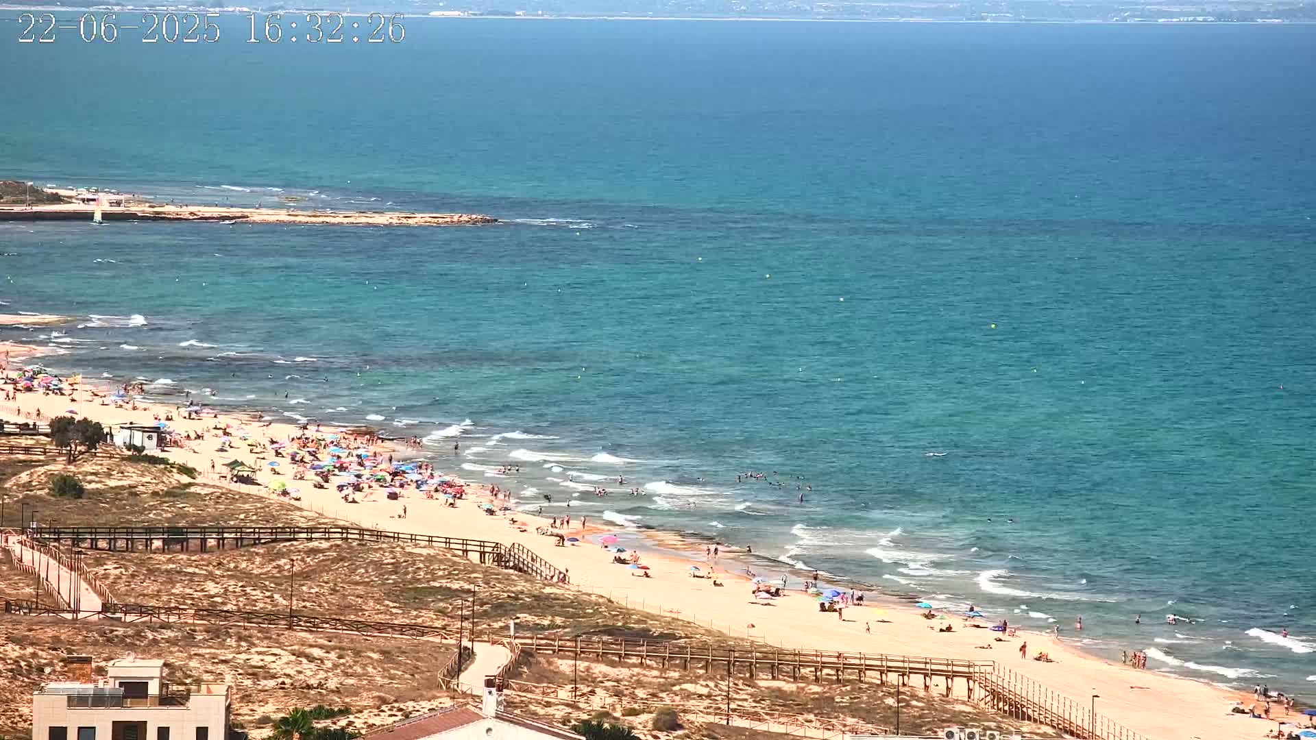A sandy beach with many people and umbrellas is bordered by a wooden walkway, overlooking a calm, teal ocean on a sunny day.