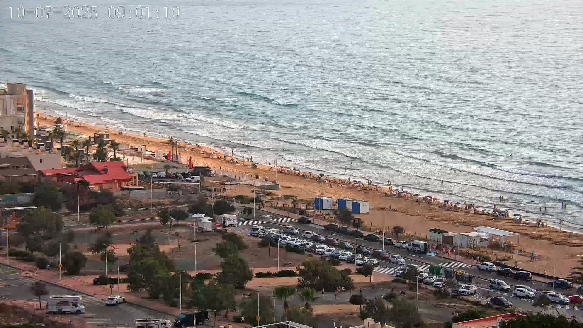 An aerial view shows a sandy beach with many people on it and in the ocean, next to a road lined with parked cars, buildings, and vegetation, under a mostly clear sky.