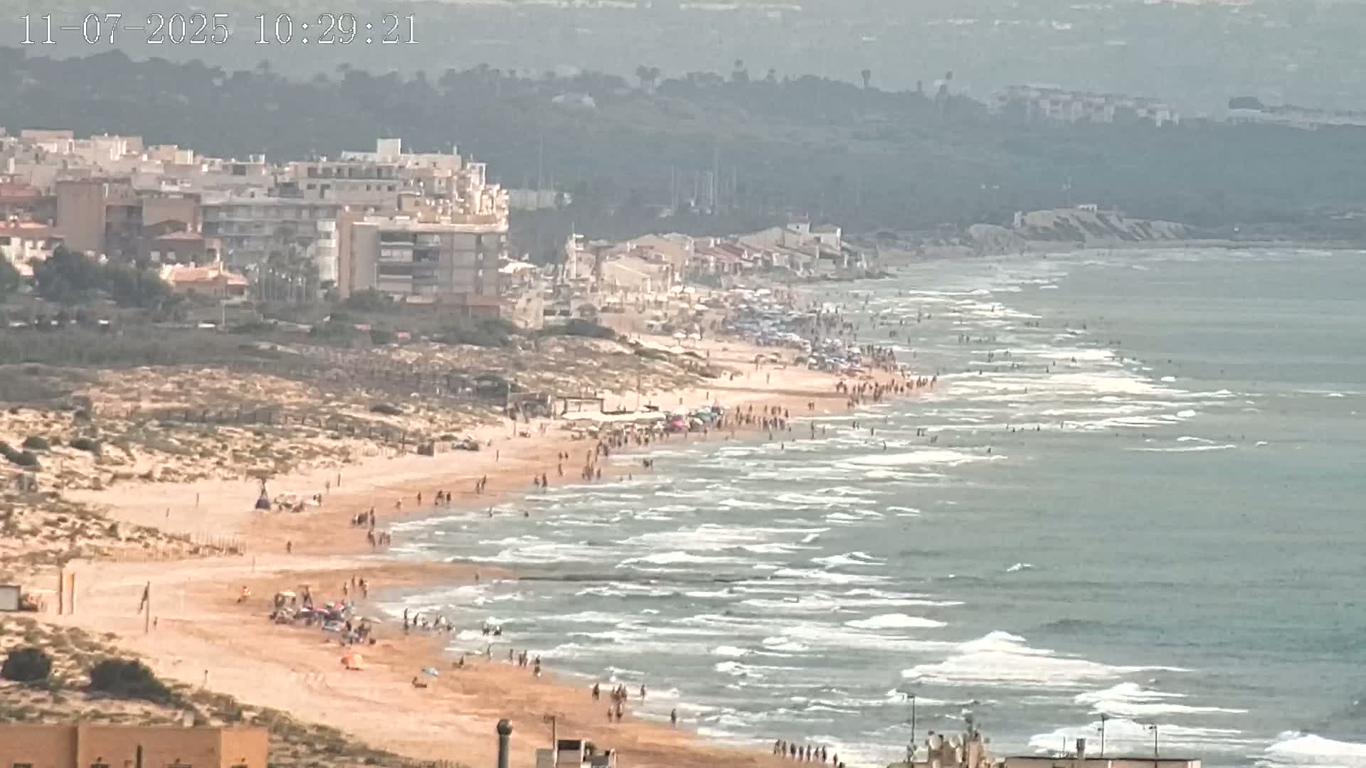A sandy beach crowded with people slopes down to a gently wavy ocean under a hazy sky.