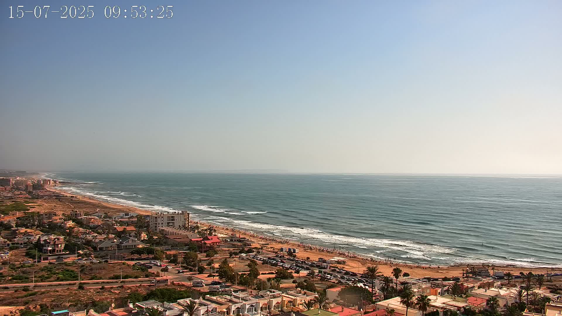An aerial view shows a beach with people on it and a town behind it, all under a clear, sunny sky.