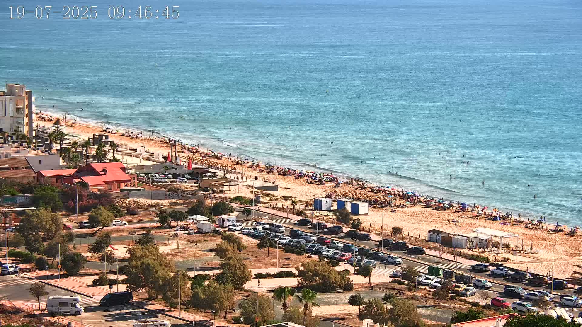 A sandy beach crowded with people and umbrellas under a sunny sky meets a calm, blue ocean.