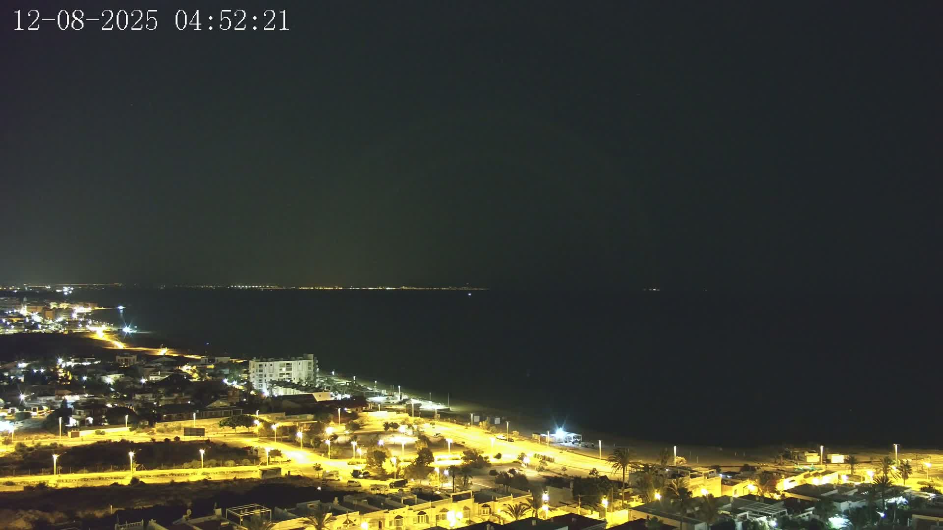 A nighttime coastal scene shows a dark sea under a clear sky, with a brightly lit town and beach stretching along the shoreline.