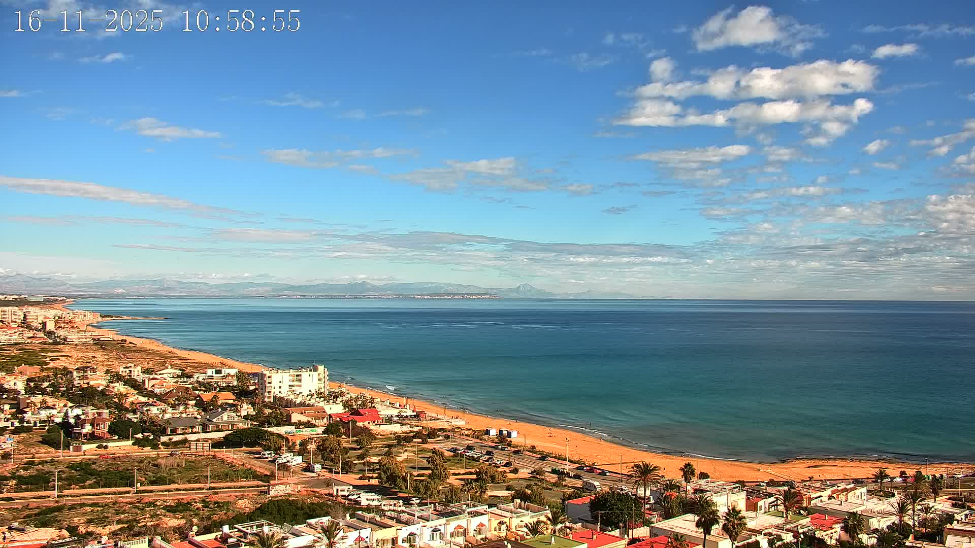 A sunny coastal landscape features a long sandy beach bordering a turquoise sea, a town with numerous buildings and palm trees stretching along the shore, and distant mountains beneath a bright blue sky with scattered white clouds.