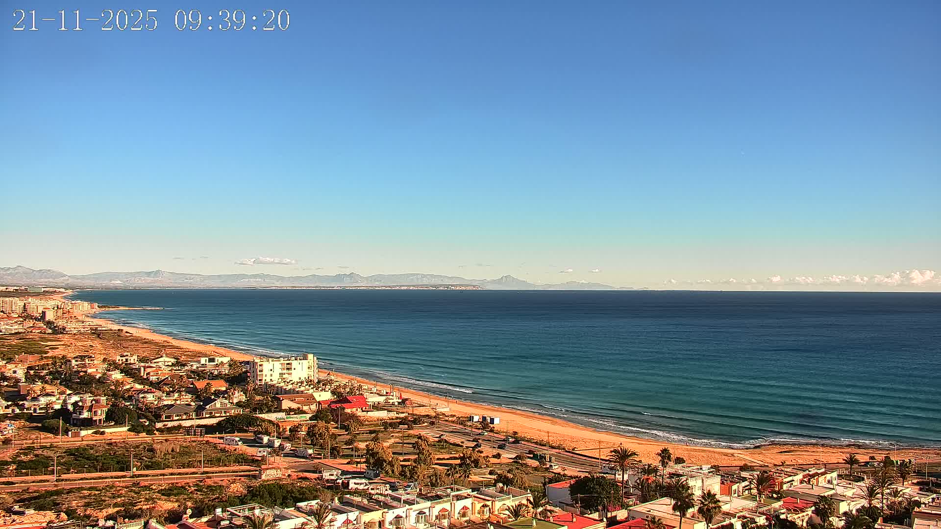 A sunny, clear day reveals a coastal town with buildings and palm trees lining a sandy beach next to a blue ocean, with distant mountains visible under a vast blue sky.