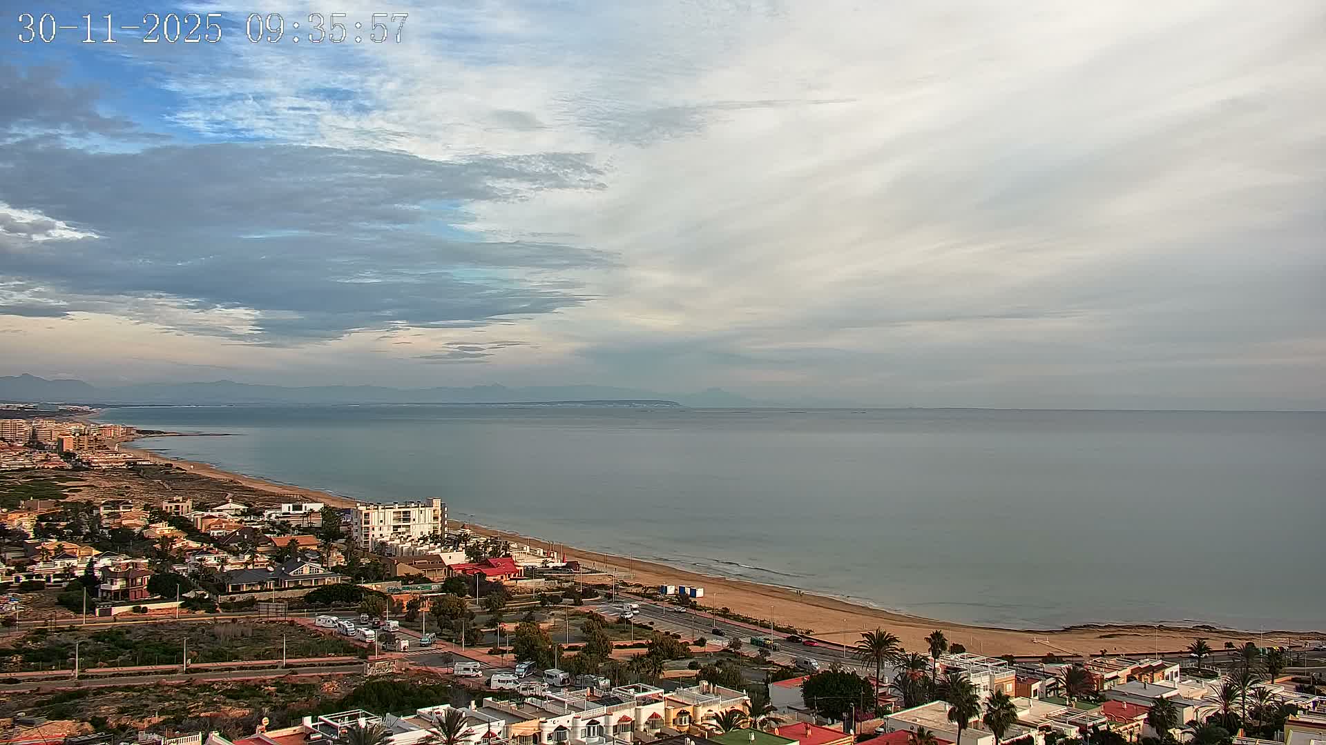 An elevated view reveals a sprawling coastal town with a sandy beach bordering a calm sea under a vast, mostly overcast sky with hints of blue, and distant mountains on the horizon.