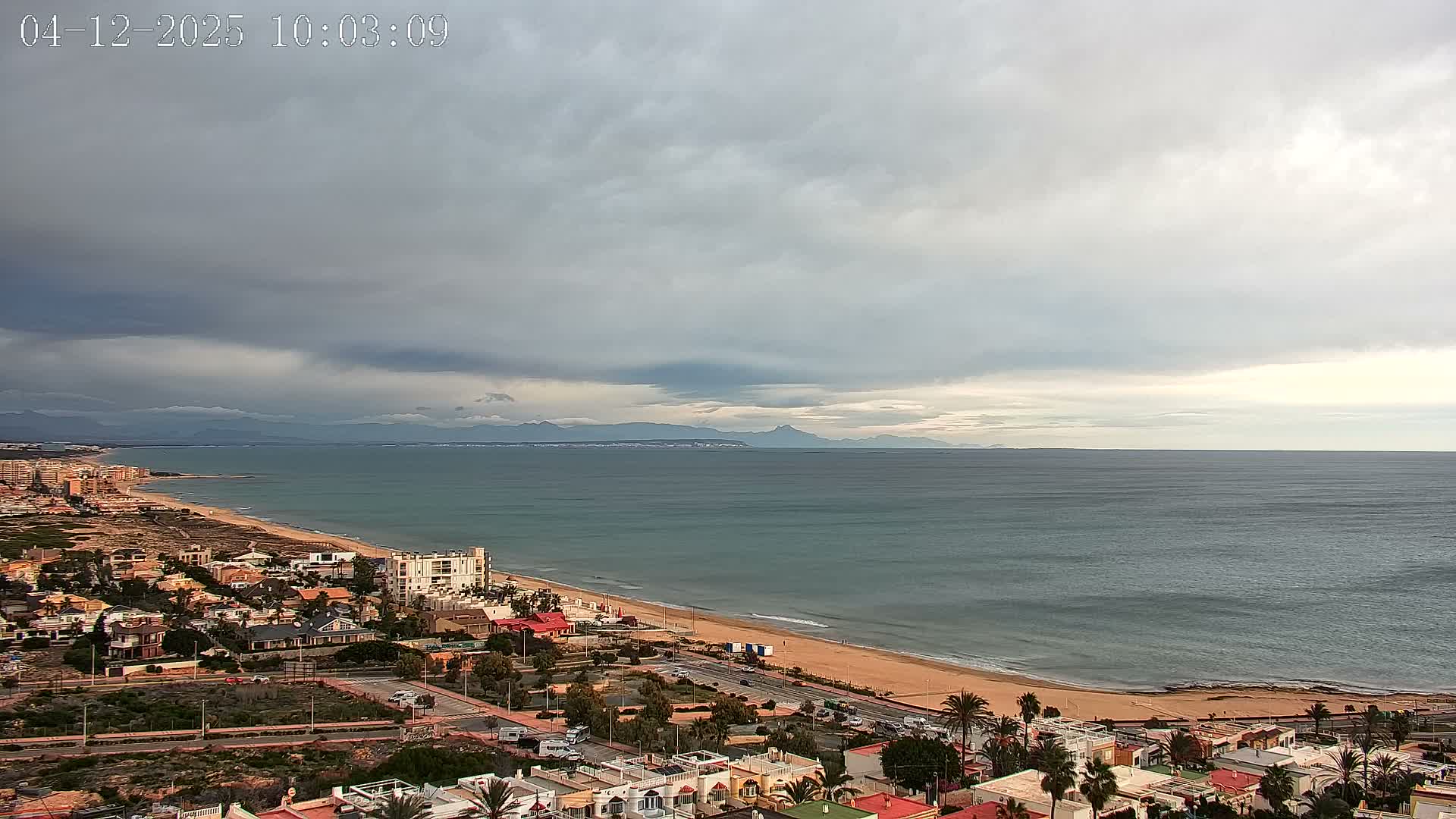 An aerial view captures a coastal town bordering a long sandy beach and the sea, with distant mountains visible on the horizon, all under a heavily cloudy and overcast sky.