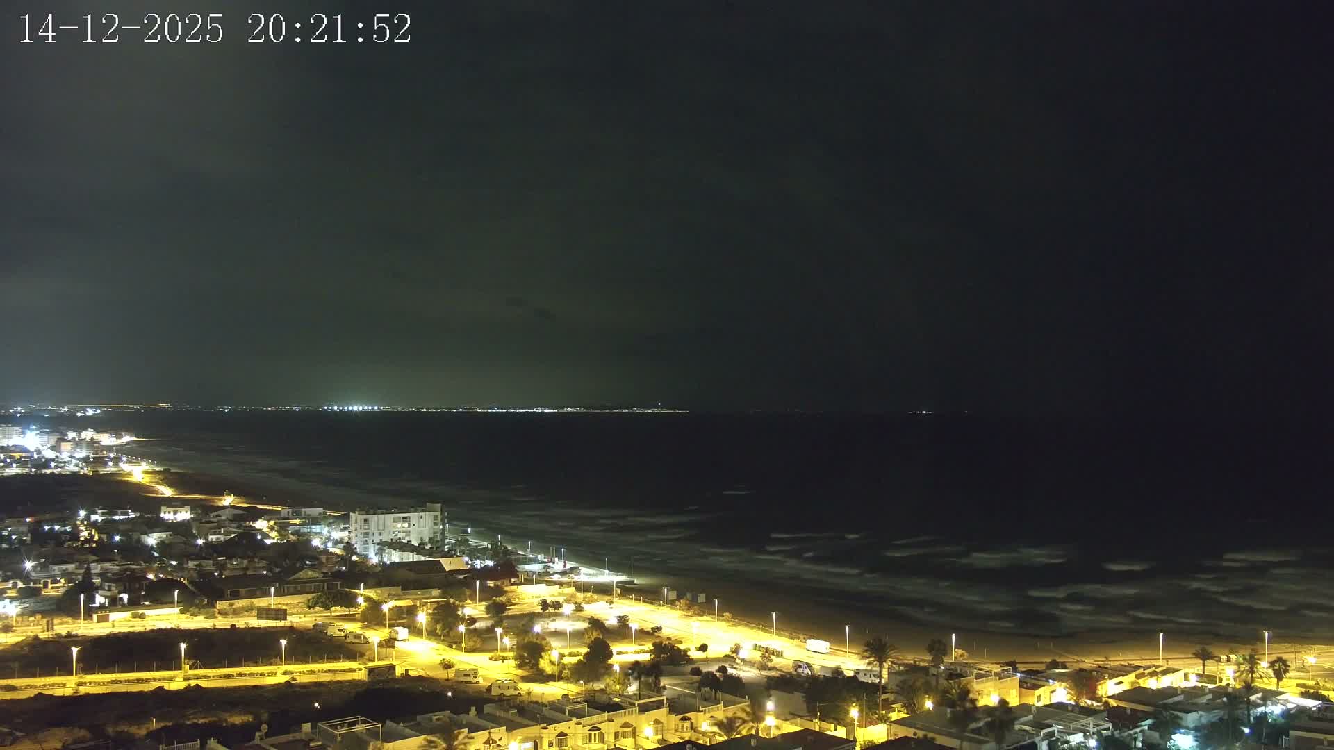 An aerial view captures a coastal town bordering a long sandy beach and the sea, with distant mountains visible on the horizon, all under a heavily cloudy and overcast sky.