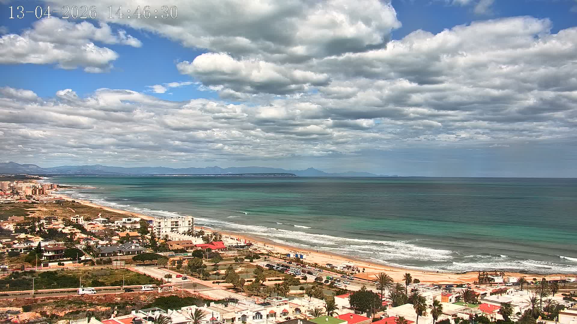 An aerial view captures a coastal town bordering a long sandy beach and the sea, with distant mountains visible on the horizon, all under a heavily cloudy and overcast sky.