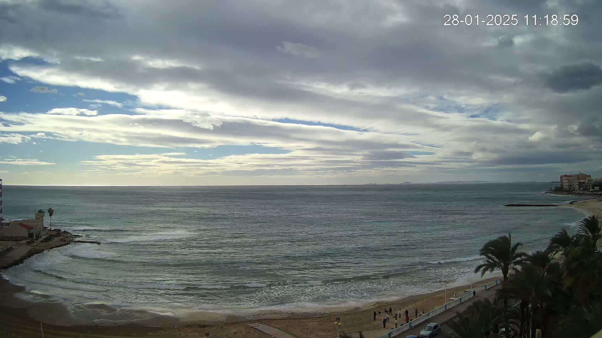 A mostly cloudy sky overlooks a calm ocean lapping a sandy beach lined with palm trees and buildings.