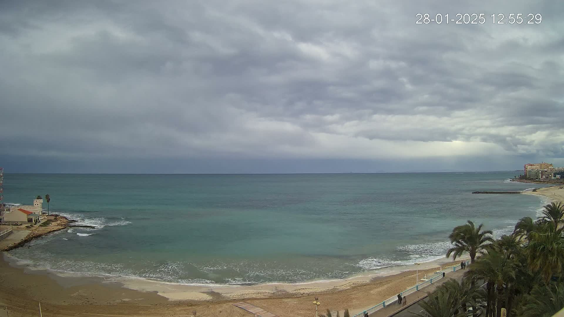 A sandy beach curves along a calm teal sea under a mostly cloudy sky.