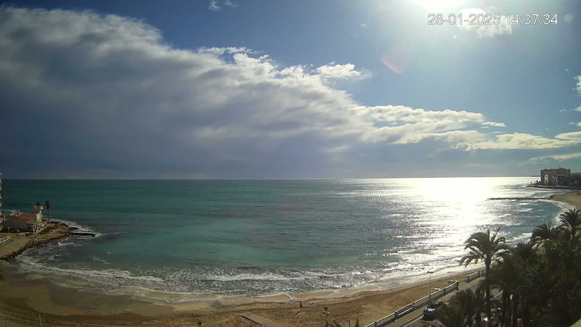 A sandy beach with calm waves and turquoise water stretches out under a partly sunny sky with fluffy clouds.