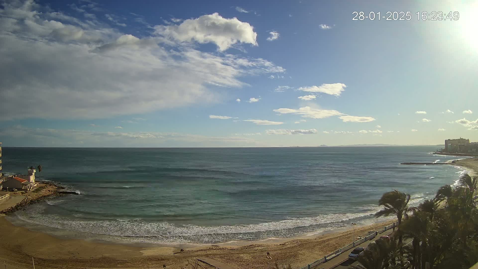 A partly cloudy day shows a sandy beach with gentle waves lapping the shore, palm trees, and buildings in the distance.