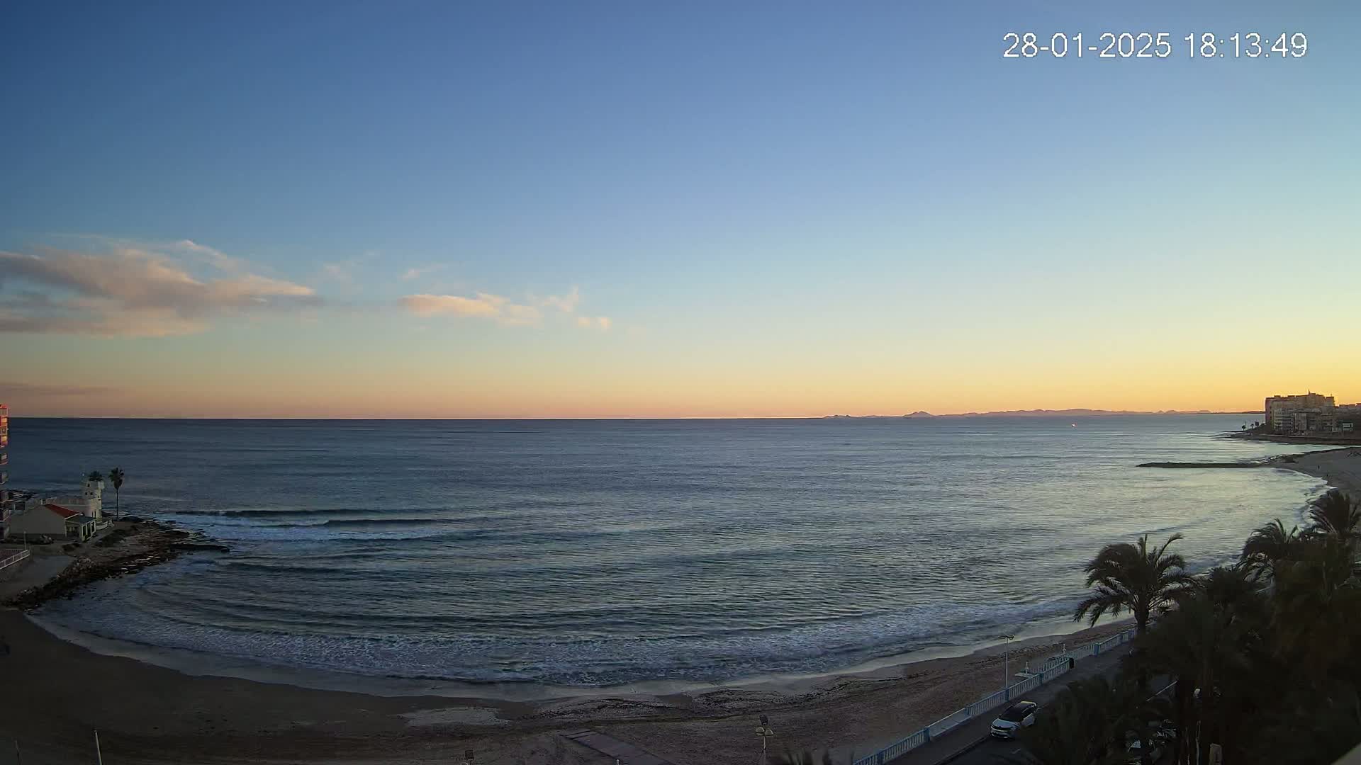 A tranquil ocean beach scene at sunset, under a mostly clear sky with a few wispy clouds.