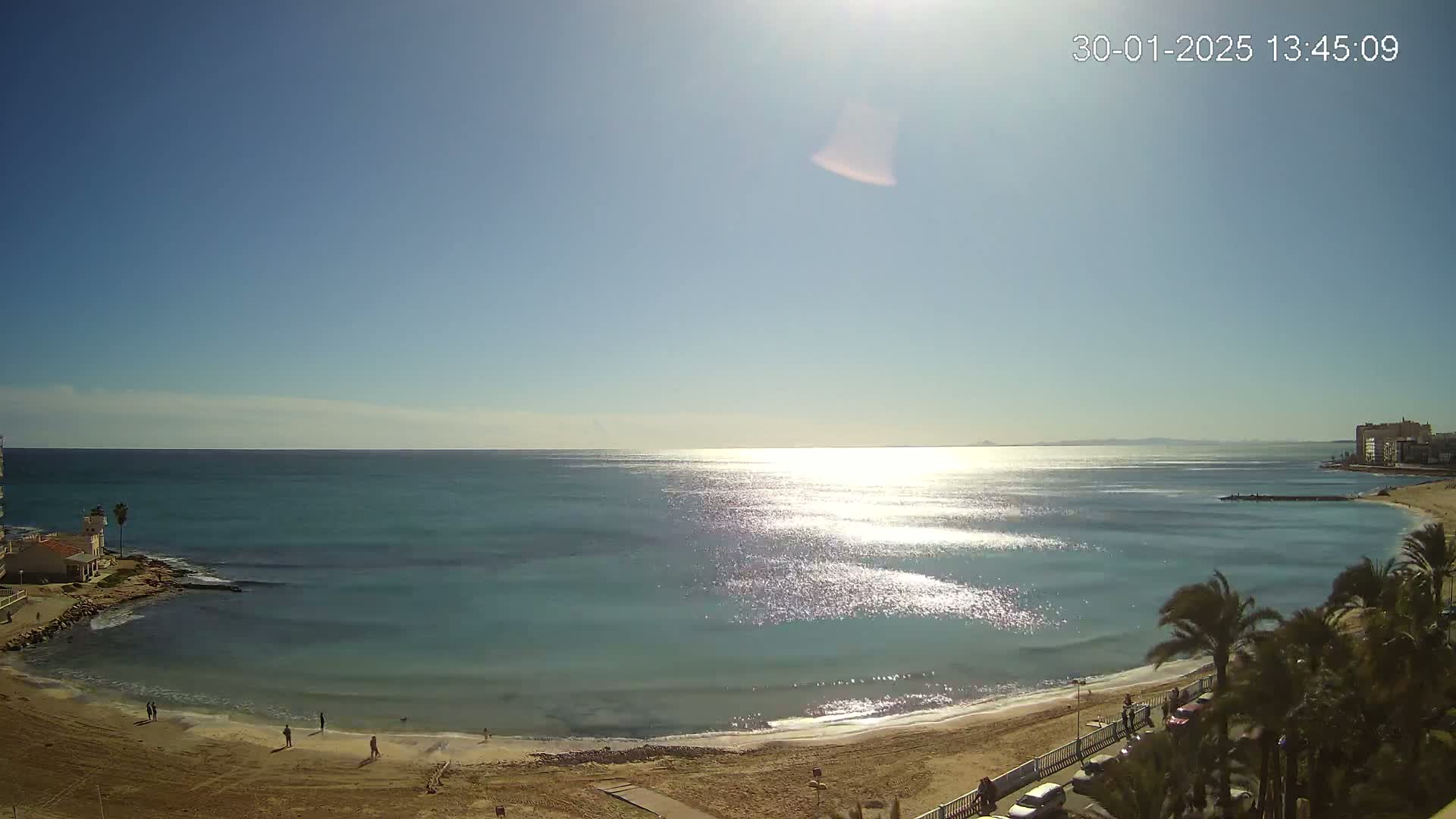A sunny day at a sandy beach with calm, sparkling turquoise water, palm trees lining the shore, and a few people on the beach.