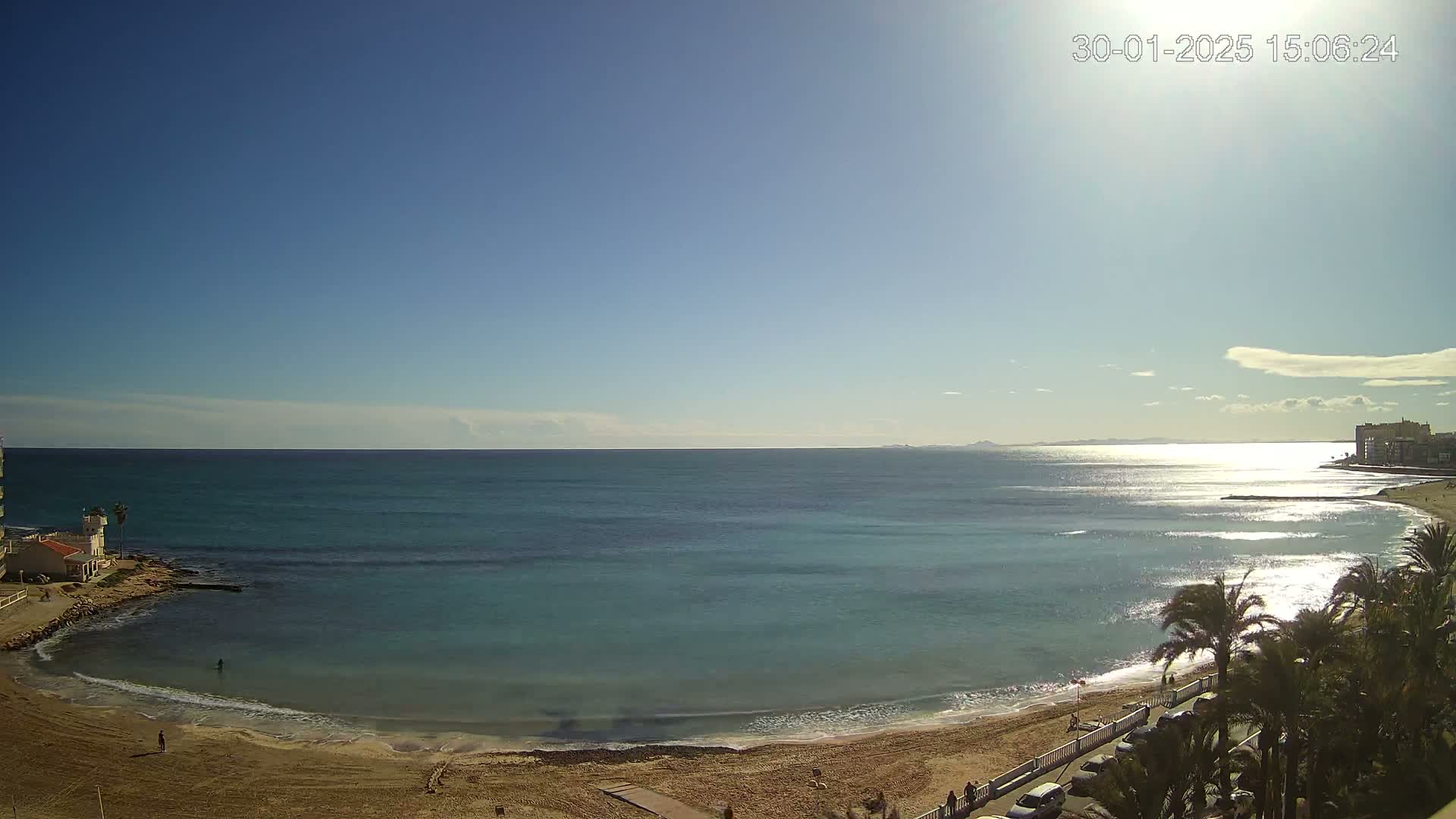 A sandy beach with calm, turquoise water stretches out under a clear, sunny sky, with palm trees and buildings visible in the background.