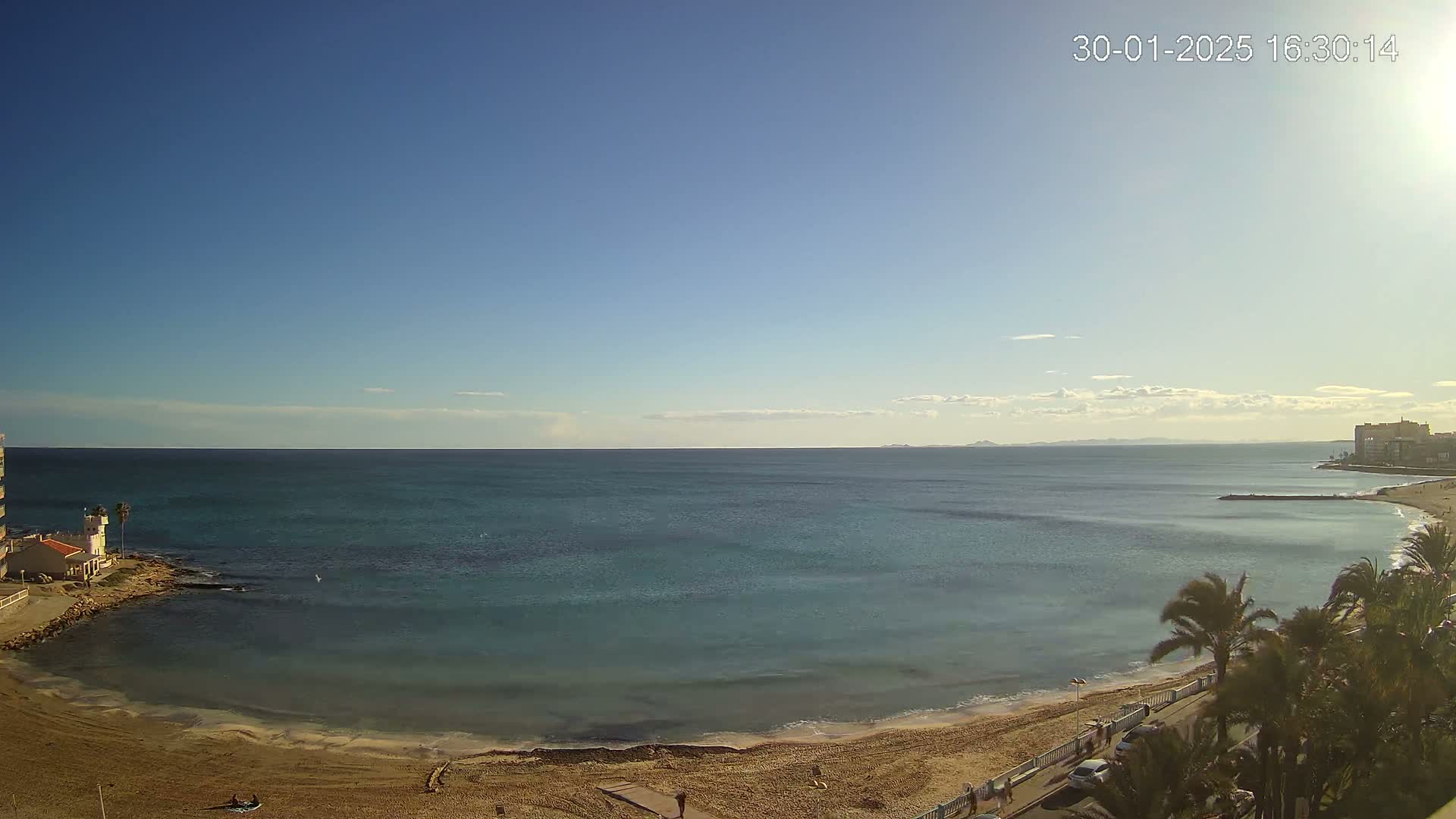 A sandy beach curves gently into a calm, teal sea under a clear, bright blue sky.