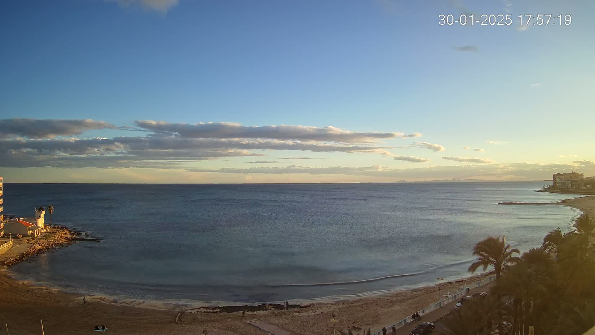 A partly cloudy sunset over a calm ocean viewed from a high vantage point, showing a sandy beach, palm trees, and buildings along the coast.