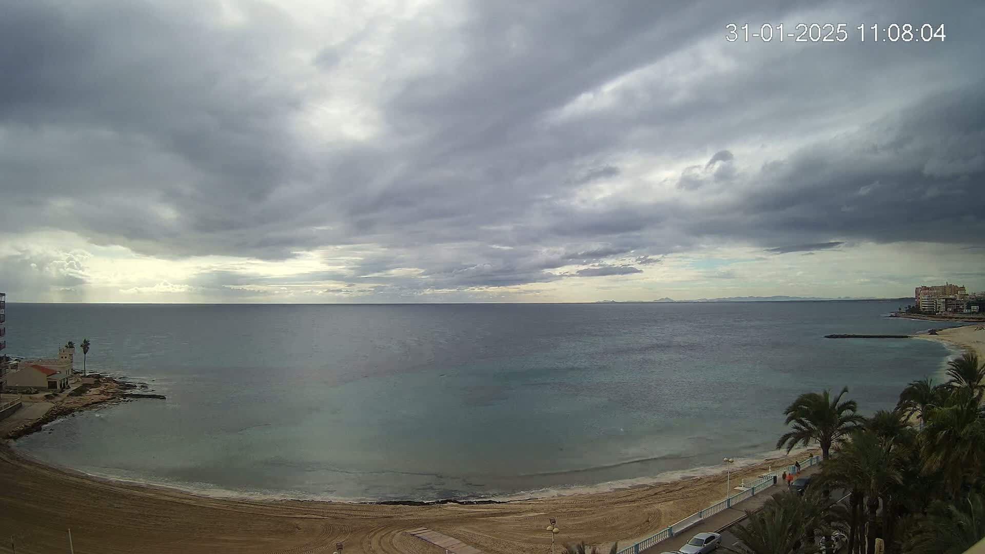 A sandy beach curves around a calm, teal-colored sea under a mostly cloudy, overcast sky.