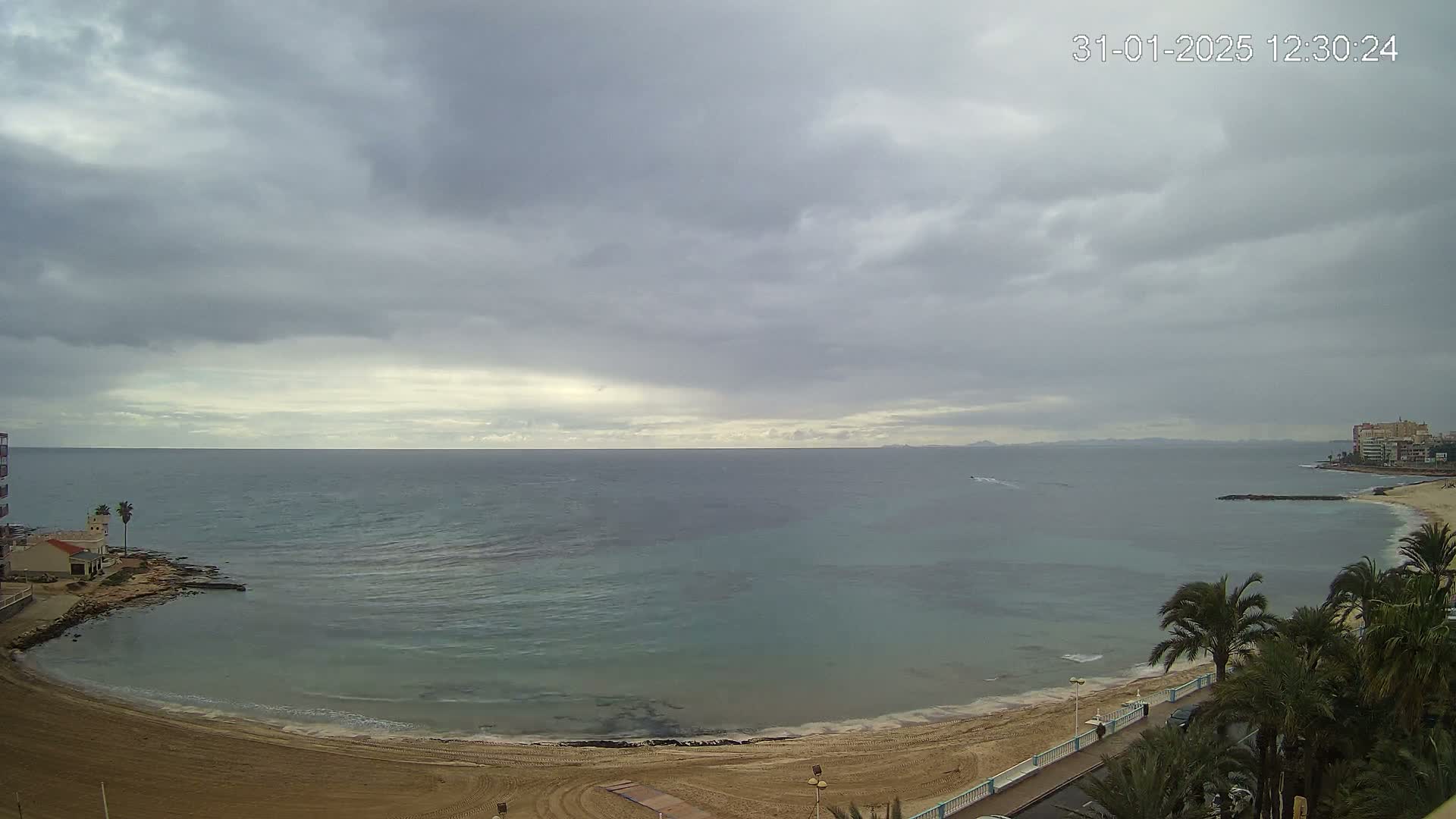A sandy beach curves around a calm, teal-colored sea under a mostly cloudy sky.