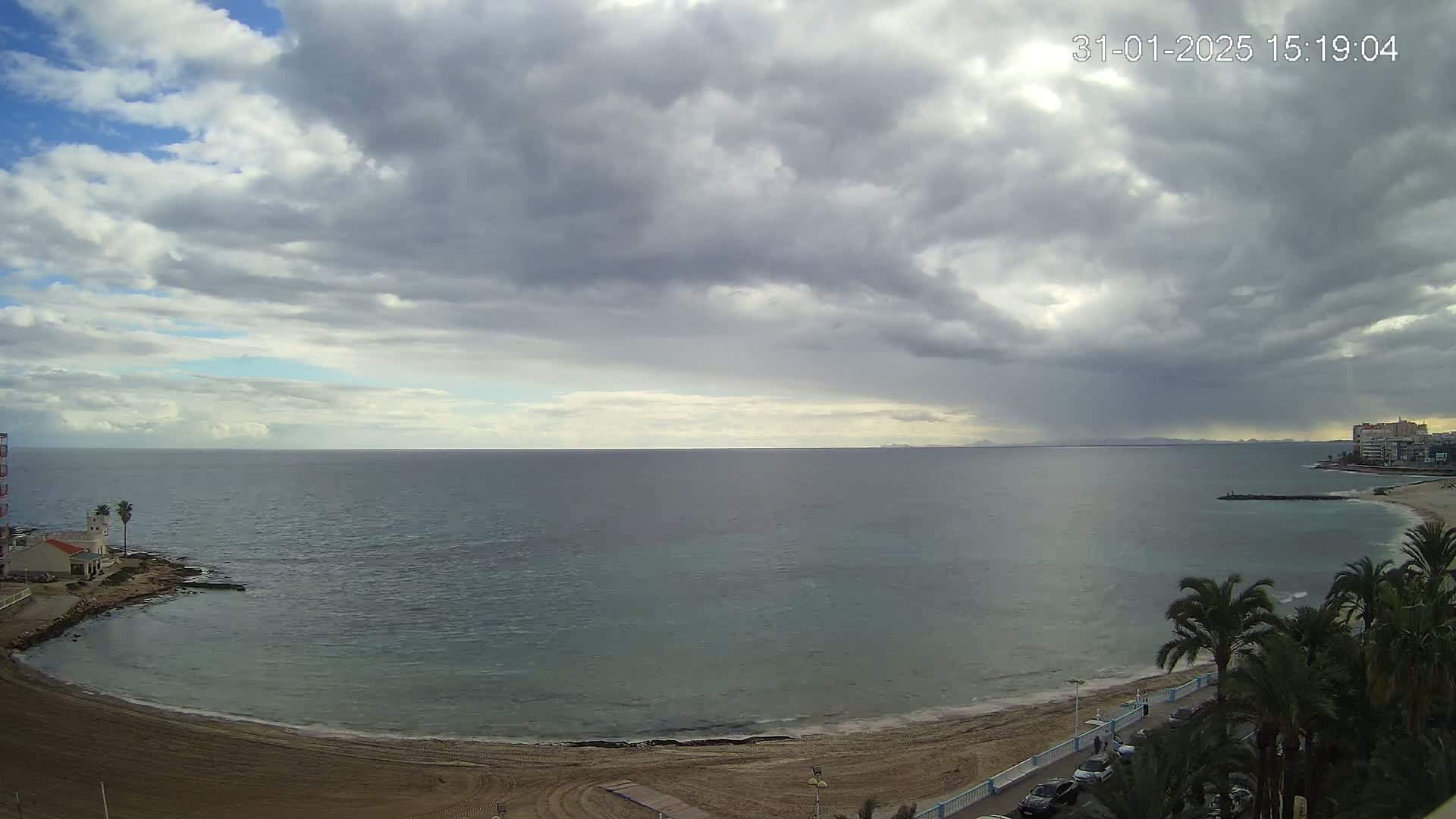 A mostly cloudy day overlooks a calm sea with a sandy beach and palm trees lining a coastal road.