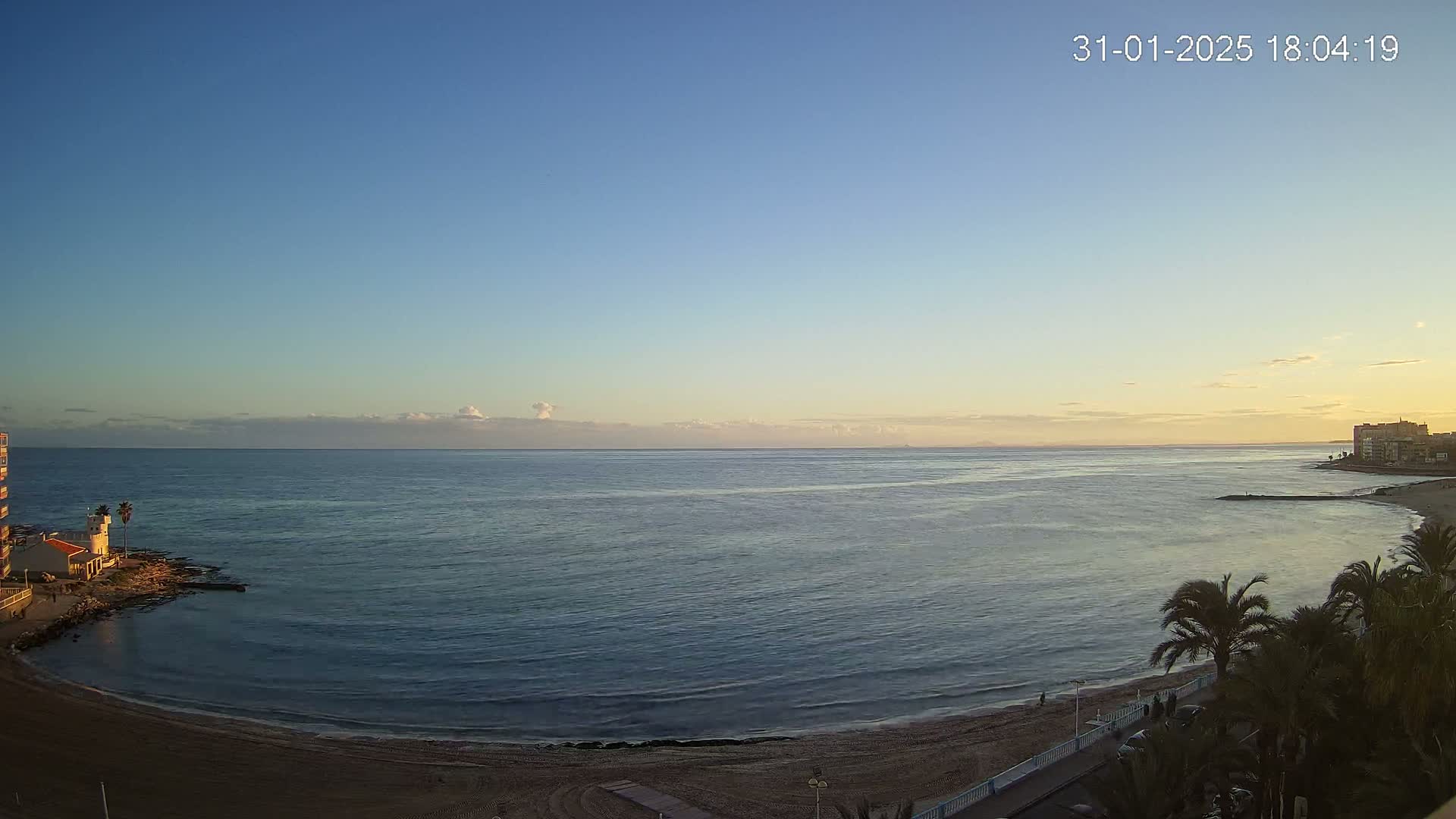 A calm ocean meets a sandy beach under a clear twilight sky, palm trees lining the shore.