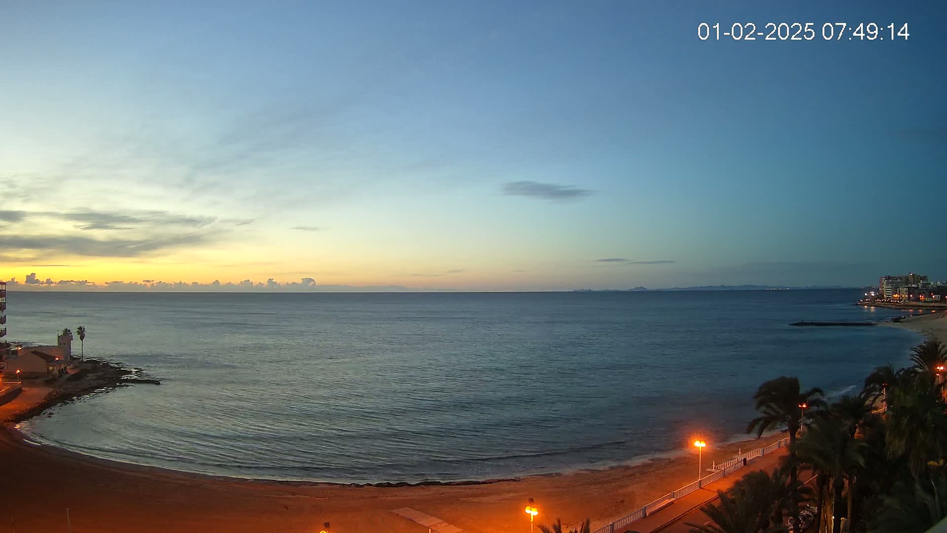 A calm ocean meets a sandy beach at sunrise, with palm trees lining a paved walkway and buildings visible in the distance.
