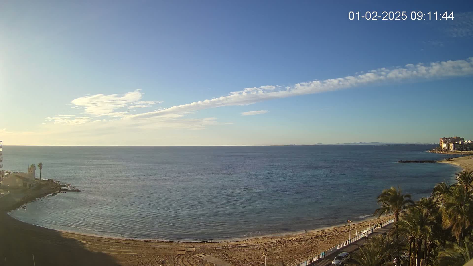 A sandy beach curves along a calm sea under a mostly clear sky with a thin line of clouds.