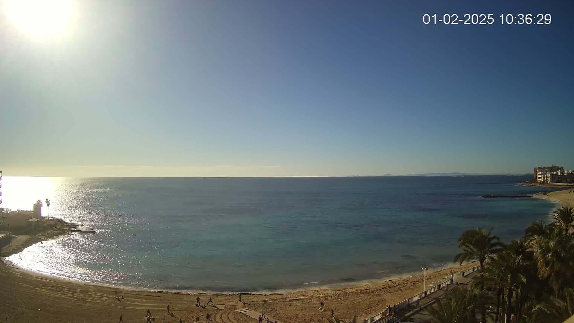 A sunny day at a beach shows a calm, turquoise sea lapping a sandy shore where people stroll; palm trees line the walkway behind the beach.